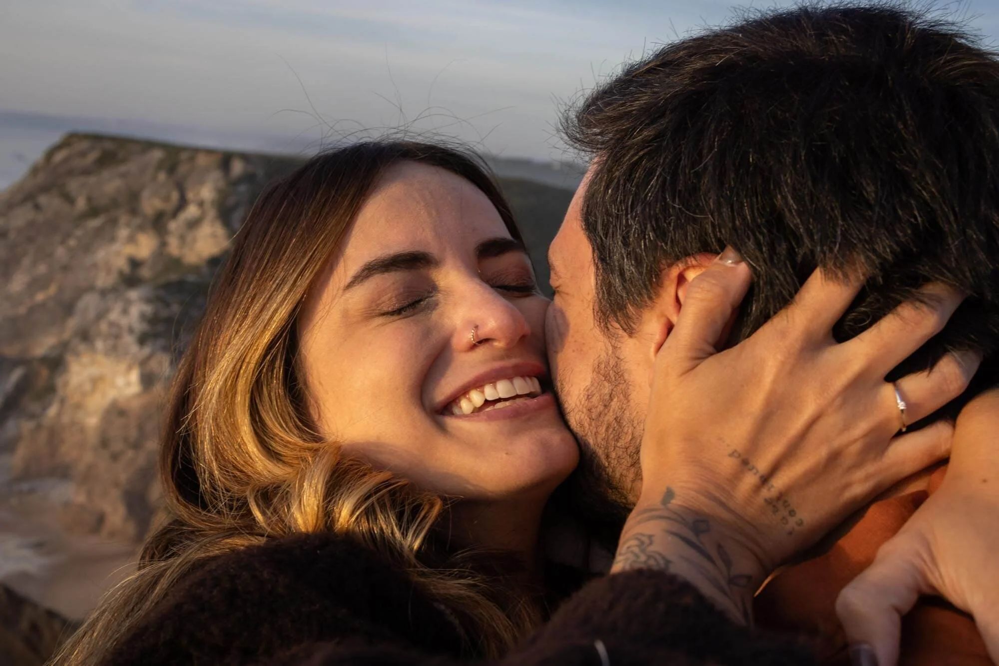 A close-up of a couple kissing outdoors near a rocky area, with the woman smiling and eyes closed.