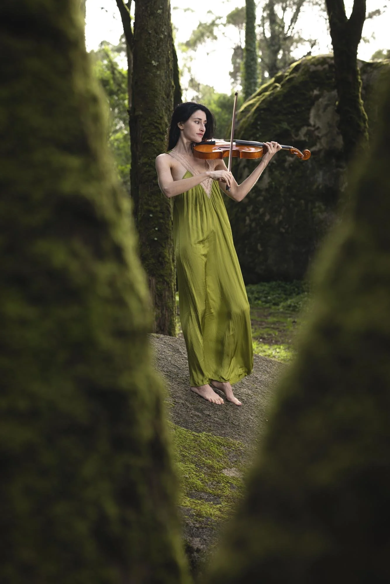 A woman in a green dress playing the violin in a moss-covered forest.
