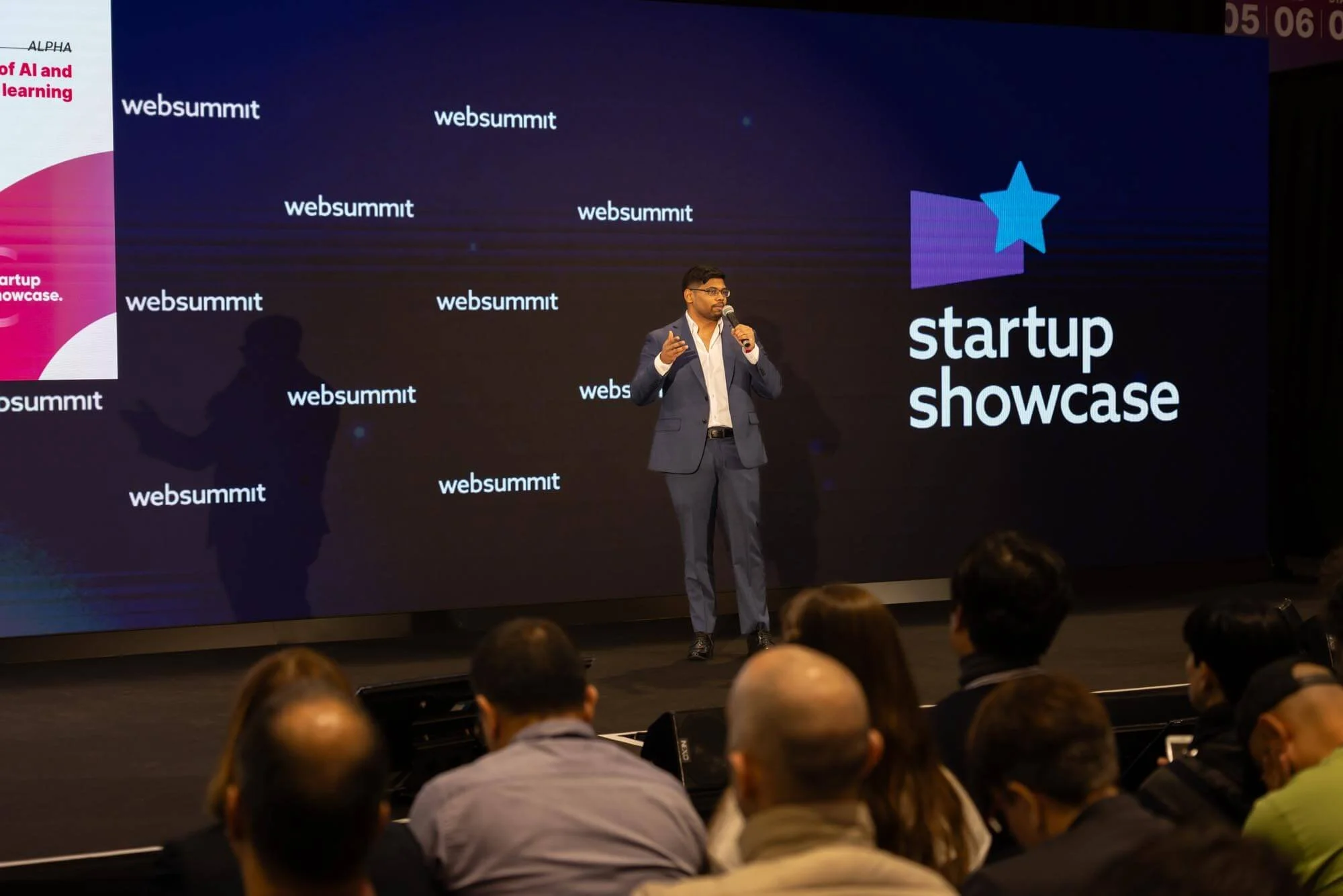A man in a suit speaking into a microphone on stage at a 'startup showcase' event, with a large screen behind him displaying the words 'websummit' repeatedly and a logo with a star and the text 'startup showcase.' Attendees are seated in front of the