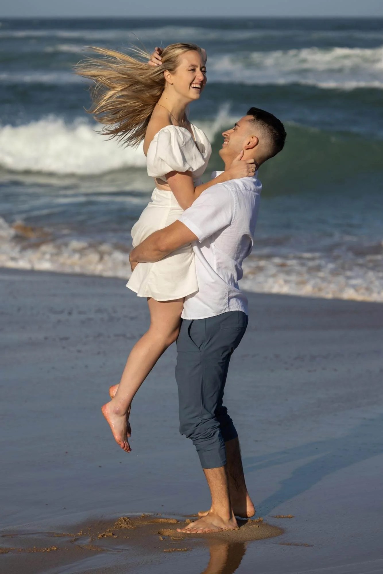 A couple with a woman in a white dress and a man in gray pants on a beach, with the man lifting the woman up while they smile at each other, waves in the background.