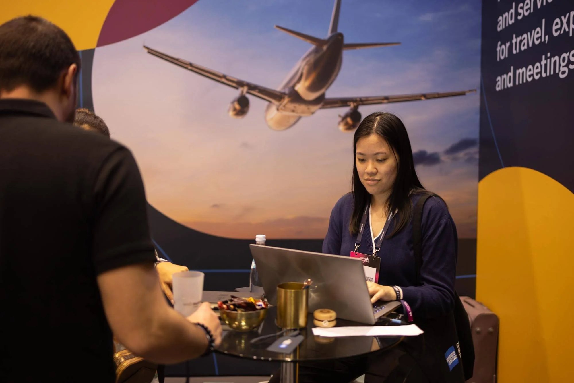 A woman sitting at a booth with a laptop in front of a large poster of an airplane flying at sunset. She is looking at her laptop with a slight smile, and there are snacks and drinks on the table.