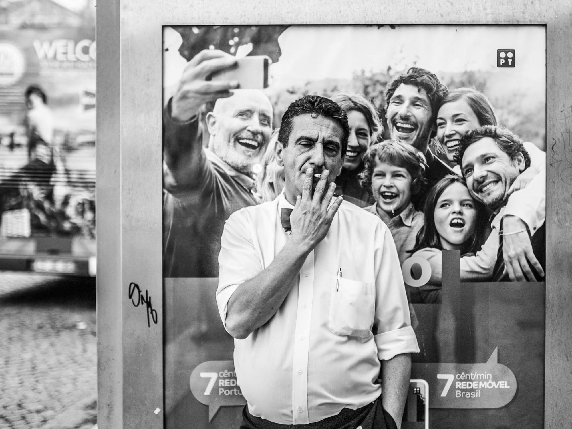 A man with a cigarette in his mouth standing in front of a large group selfie in black and white.