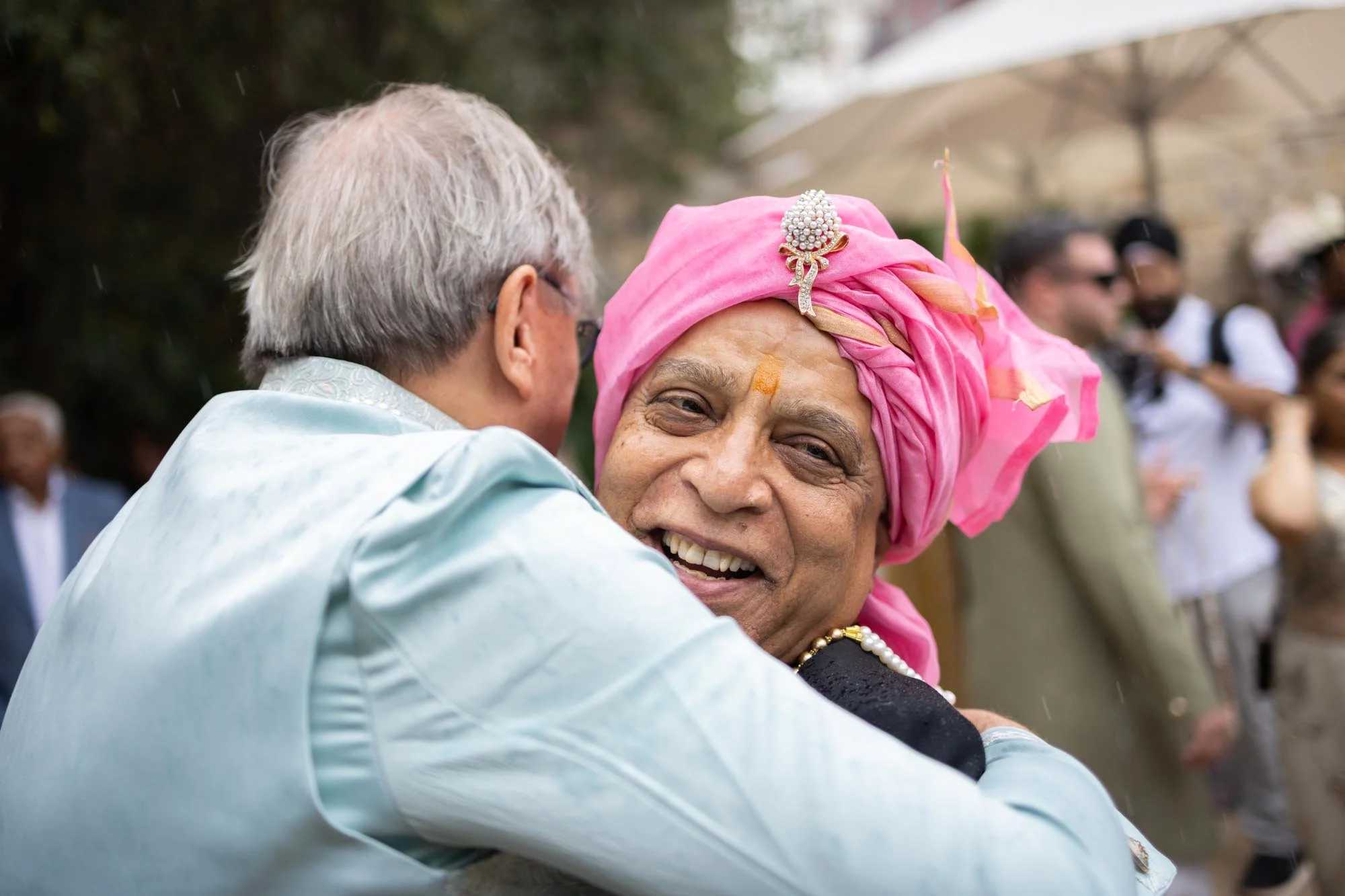 Two men hugging at an outdoor event, one wearing a pink turban and traditional attire, the other in a light-colored shirt, with people in the background.
