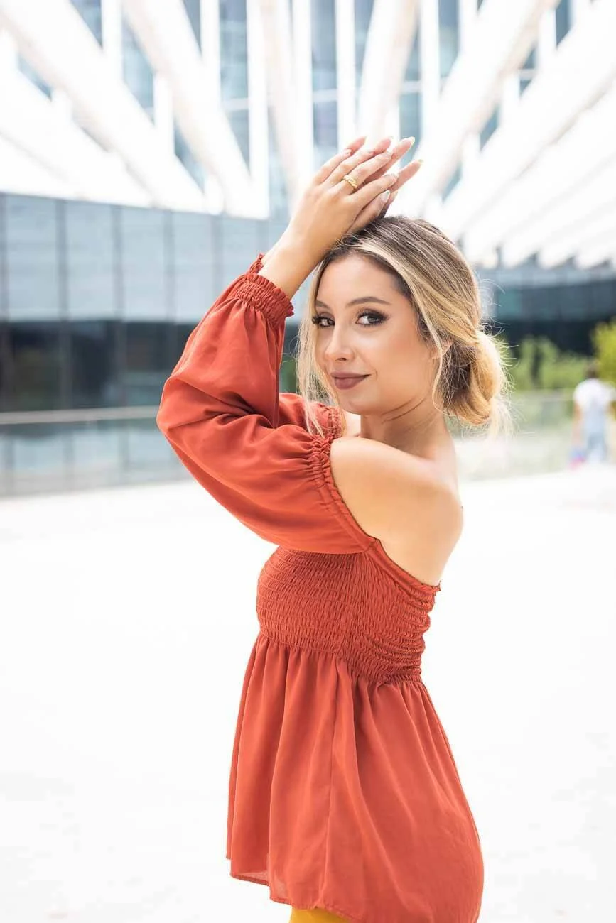 Young woman with blonde hair, wearing a red off-shoulder dress, posing outdoors in front of modern glass buildings.