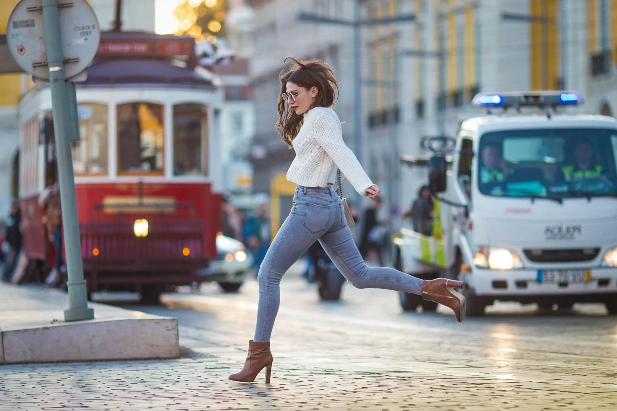 A woman wearing a white sweater, gray jeans, and brown high-heeled boots jumping on a city street, with a streetcar and cars in the background.