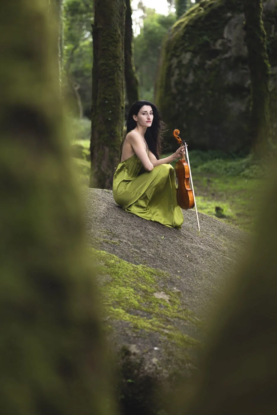 A woman with black hair in a green dress sitting on a mossy rock in a forest, holding a violin and bow.