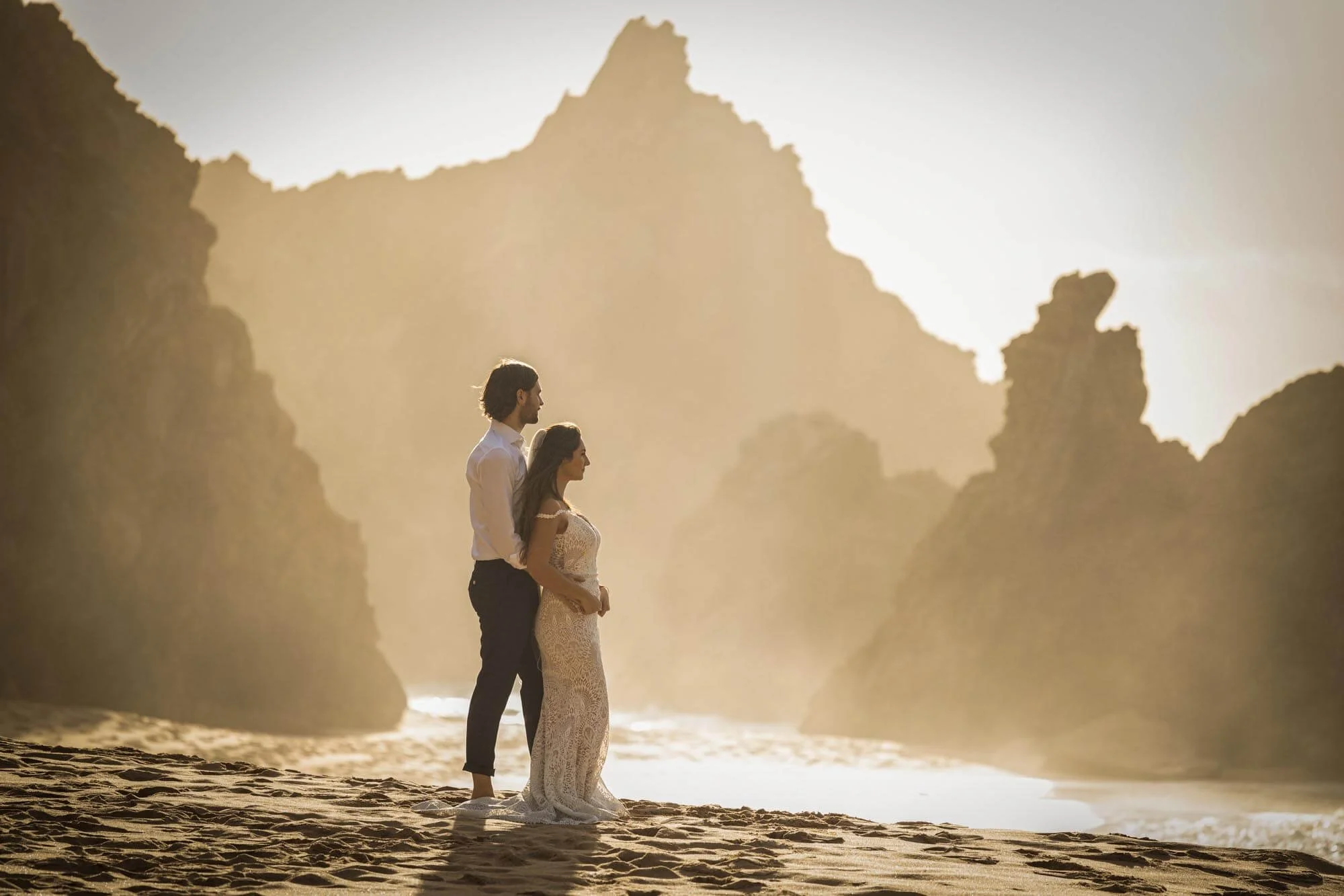 A couple in wedding attire standing on a sandy beach with large rocky formations in the background during sunset or sunrise.