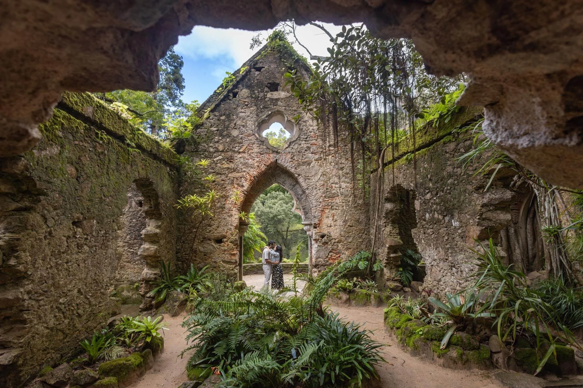 A couple hugging inside the ruins of an old stone building surrounded by lush green plants and trees.