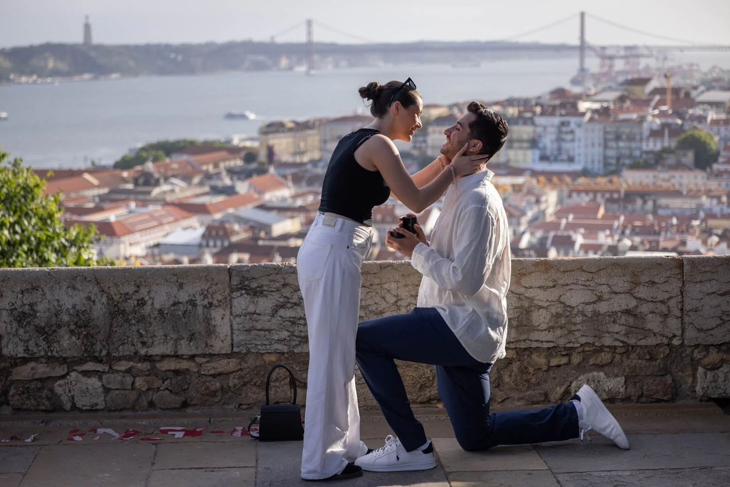 A man is proposing marriage to a woman on a scenic overlook with a city and a body of water in the background