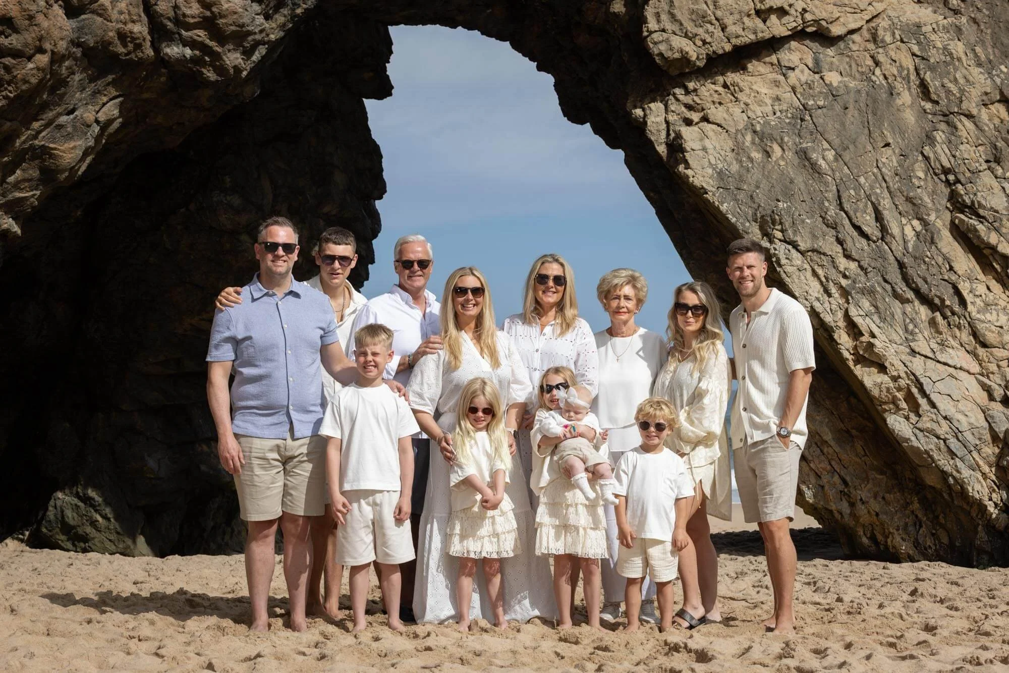 A large family group of fifteen people posing on a sandy beach in front of a natural rock arch formation. They are all dressed in white or light-colored clothing, some wearing sunglasses, smiling and standing closely together.