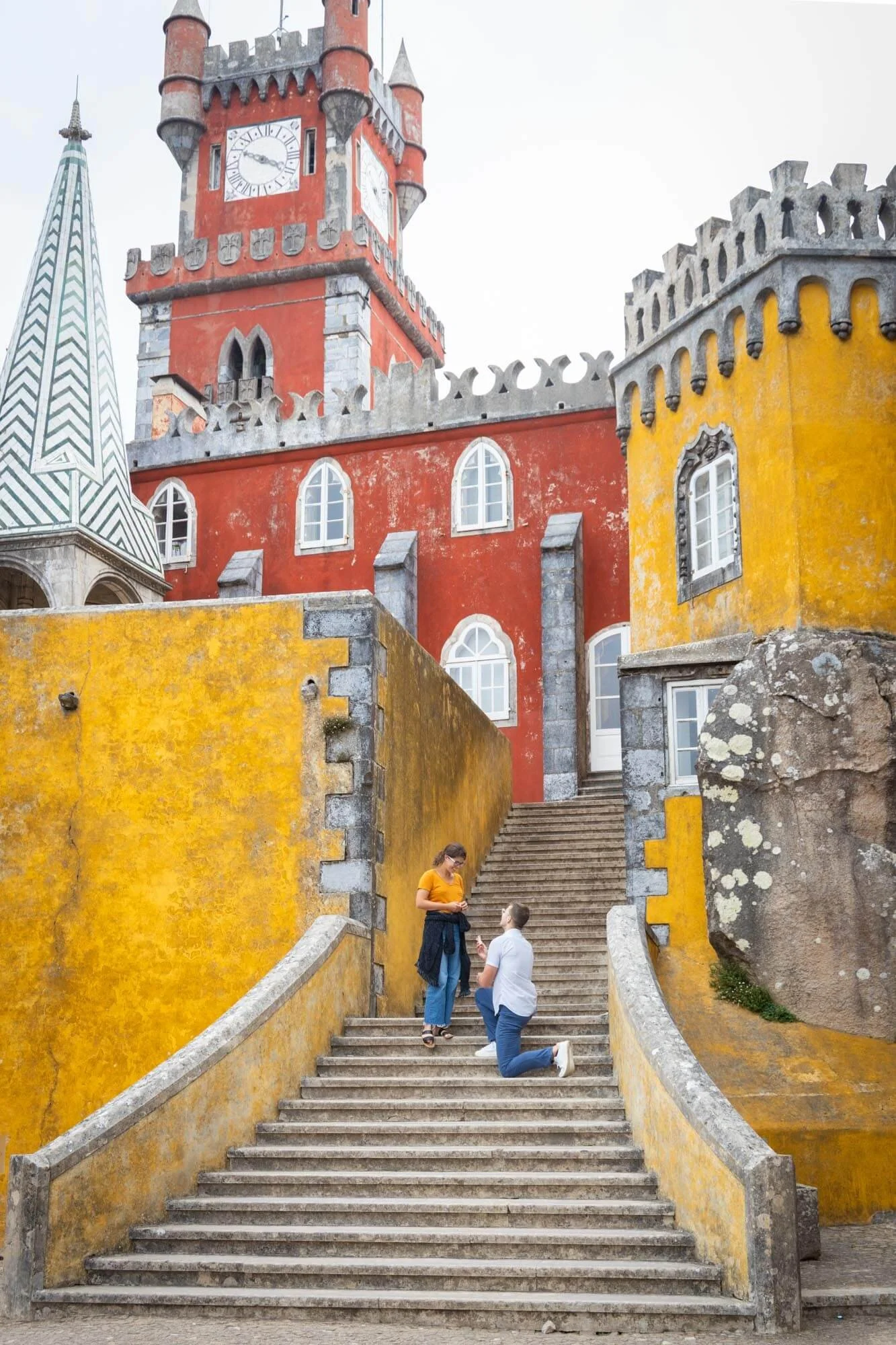 A couple on a staircase in front of colorful castle-like buildings with red, yellow, and white walls and clock tower.