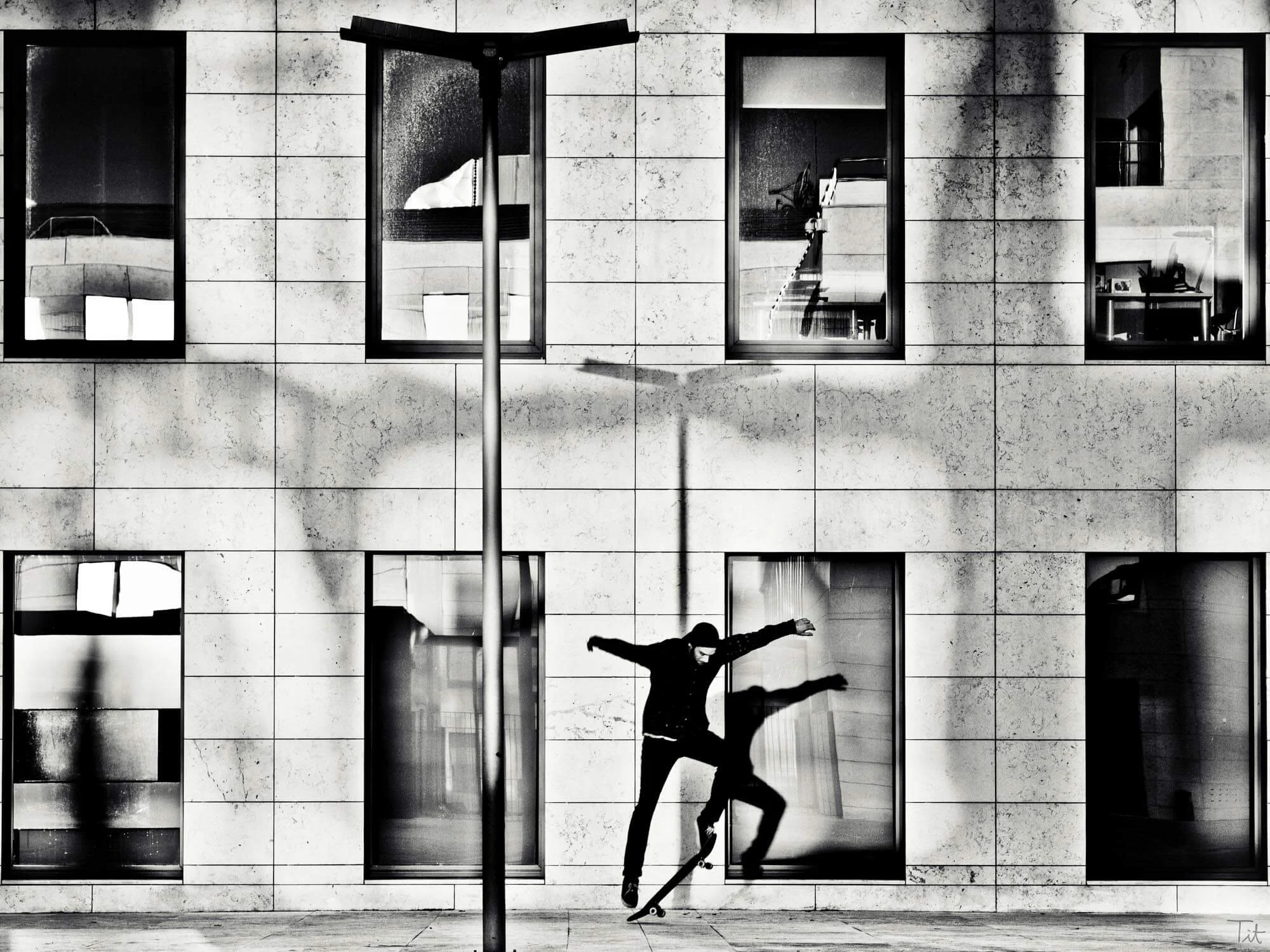 A skateboarder performing a trick in front of a modern building with large windows at night. The building's exterior is lit, creating reflections on the glass, and a streetlight casts a shadow on the wall.