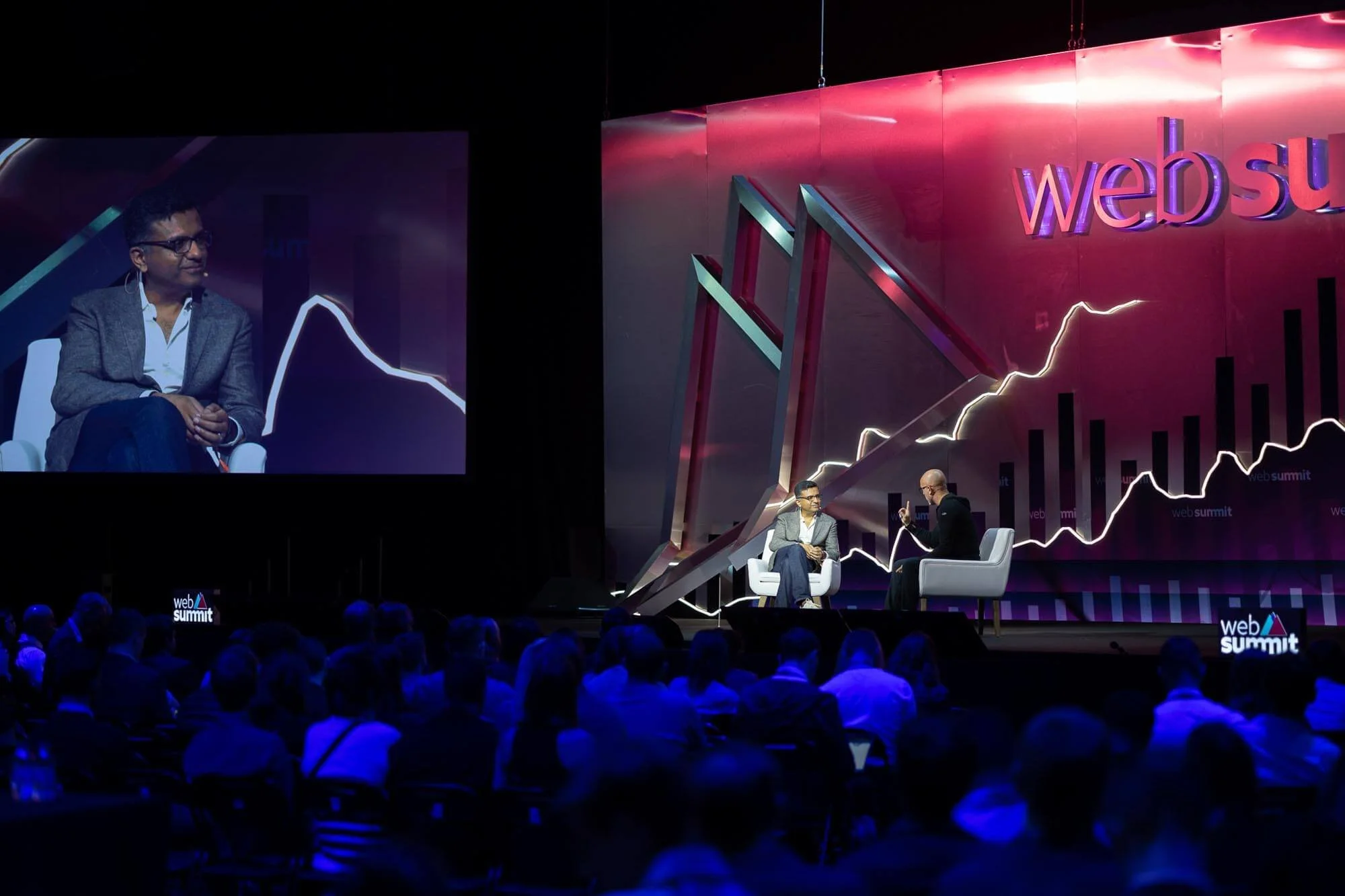 A man and woman are engaged in an interview on stage at a web summit, with a large screen and graph lines in the background and an audience watching.