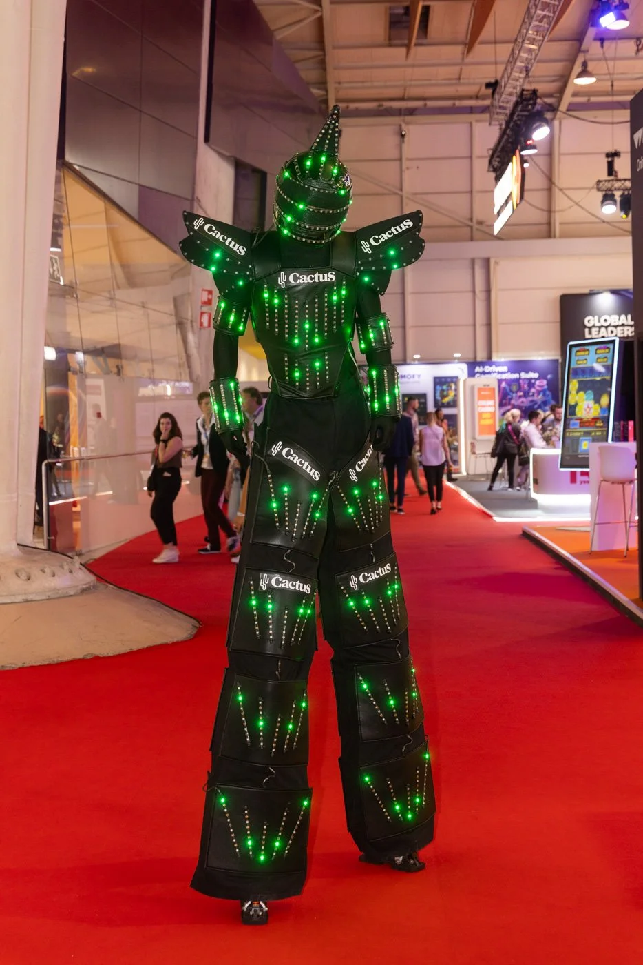 A person in an intricate costume adorned with green LED lights, resembling a robotic cactus, at an indoor exhibition with booths and attendees.