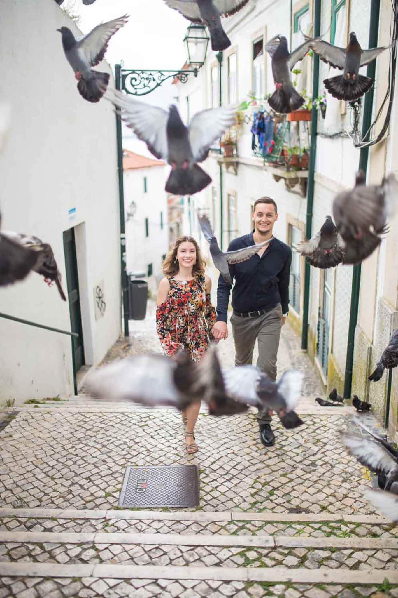 A young couple walking hand in hand down a cobblestone alleyway of Alfama, surrounded by flying pigeons, with the woman wearing a floral dress and the man in a dark shirt and khakis.