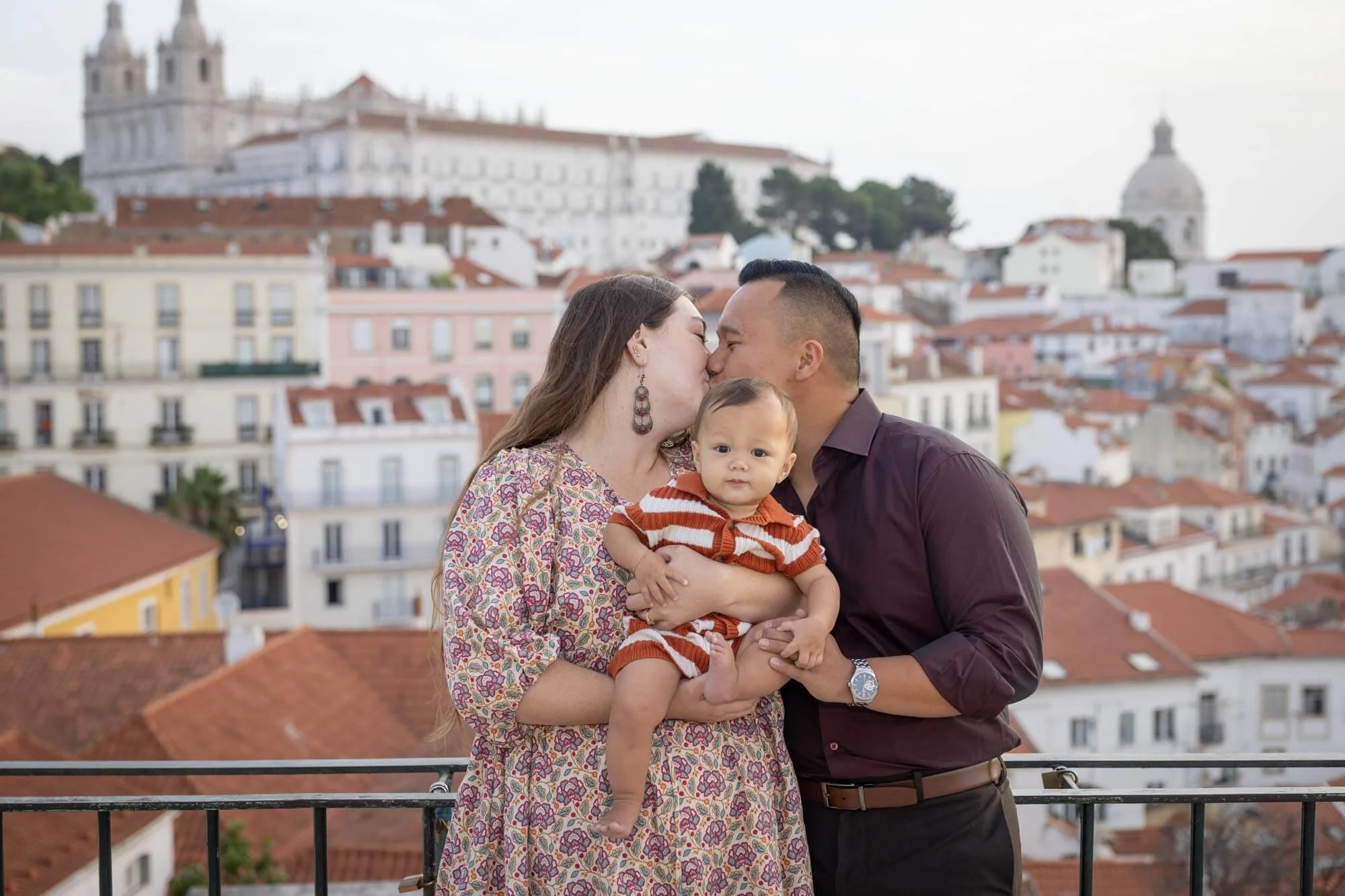 Family photo session in Alfama, a couple kissing and holding a child in the middle with old city of Alfama in the background.