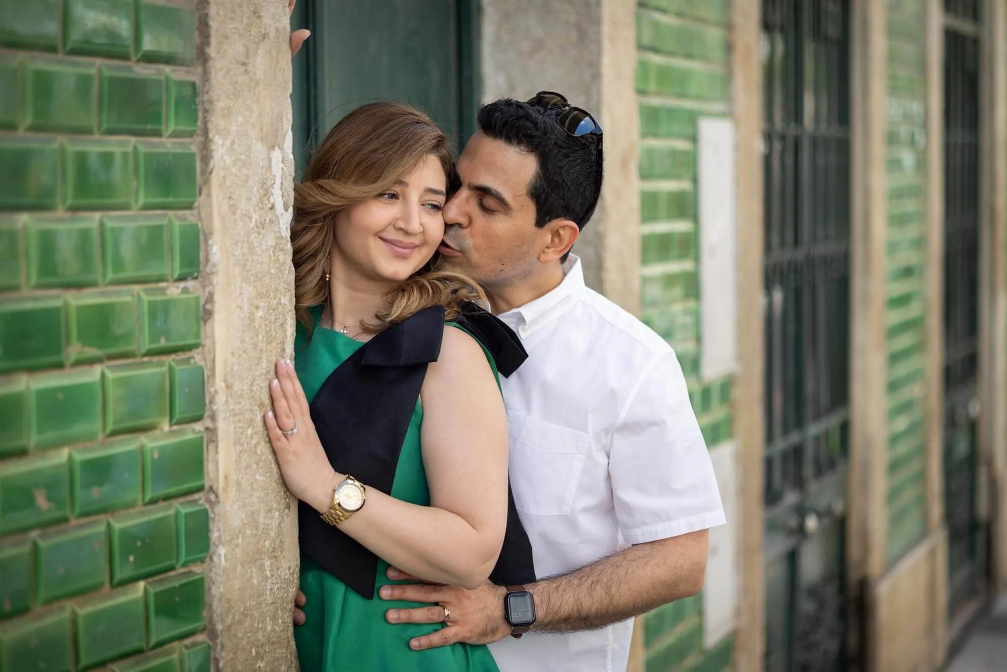 A couple embraces and shares an intimate moment against a green-tiled wall. The woman is smiling softly, wearing a green dress with black accents, a gold watch, and a ring. The man is kissing her cheek, dressed in a white shirt, and wearing a smartwa
