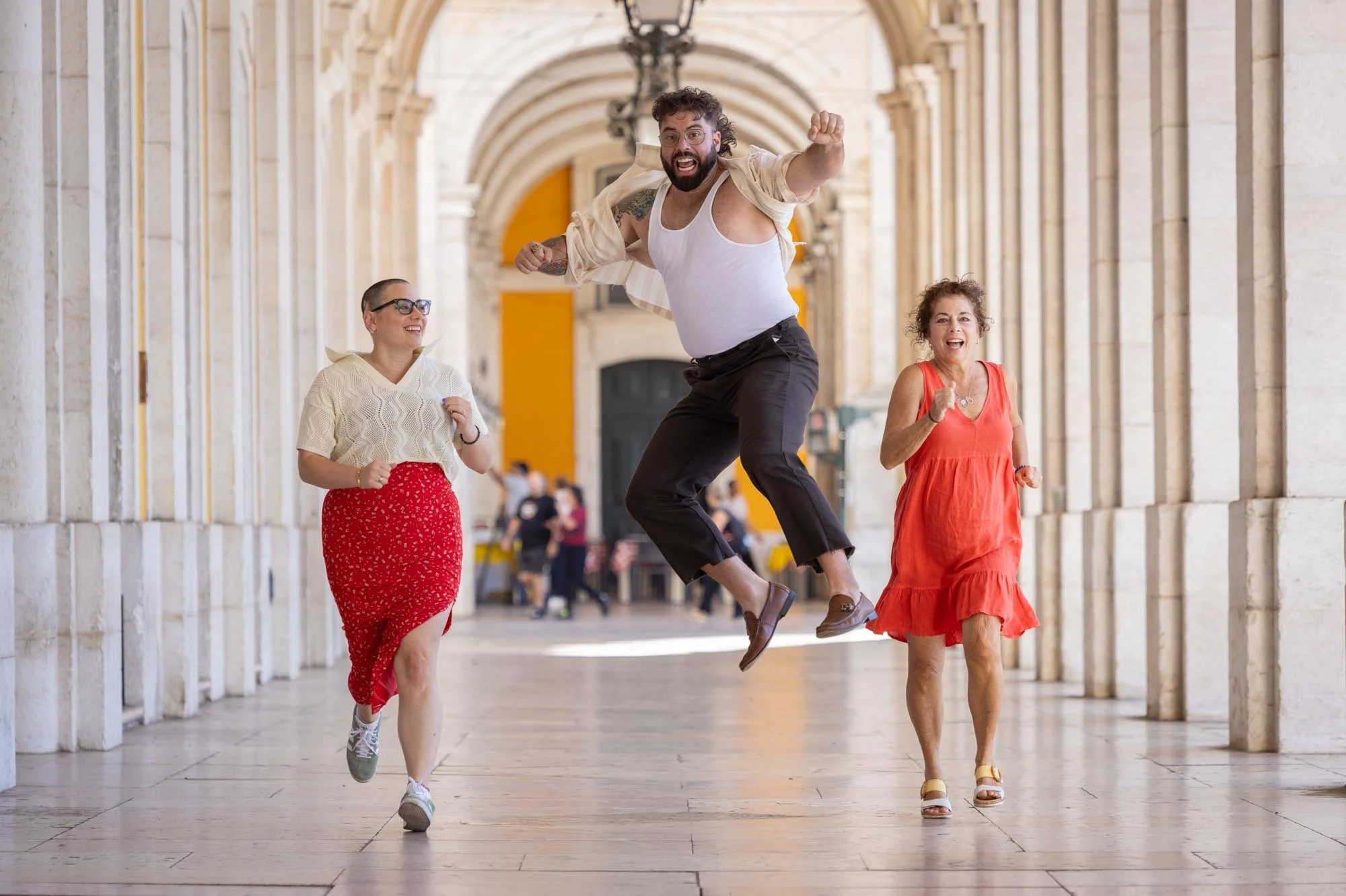 Three joyful women and one man running and jumping in a historic archway corridor with stone columns, during the daytime.