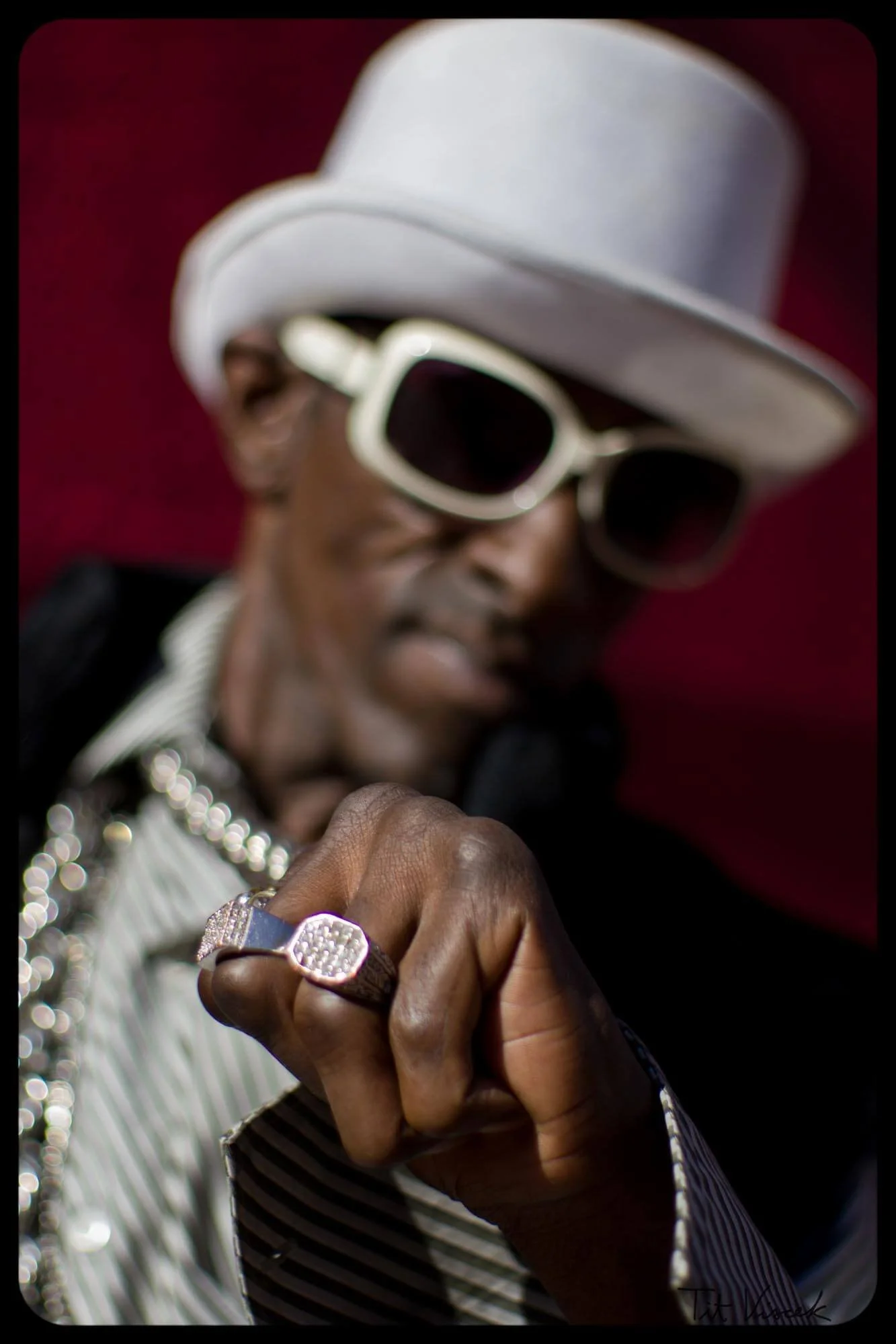 A man wearing a white fedora hat, white sunglasses, a striped shirt, gold chains, and a large ring, pointing at the camera with a confident expression.