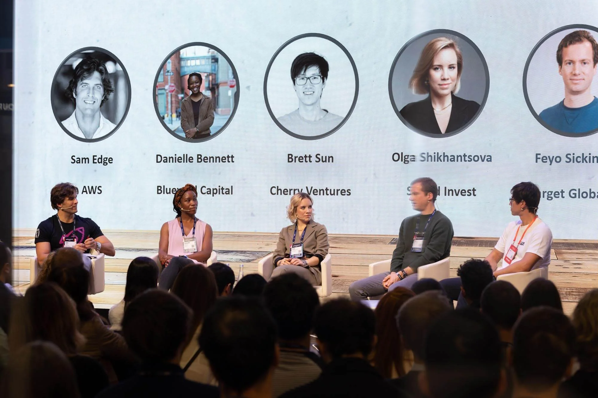 Panel discussion at a conference with five speakers sitting on stage, and a large screen behind displaying their photos and names, including Sam Edge, Danielle Bennett, Brett Sun, Olga Shikhantsova, and Feyo Sickinga.