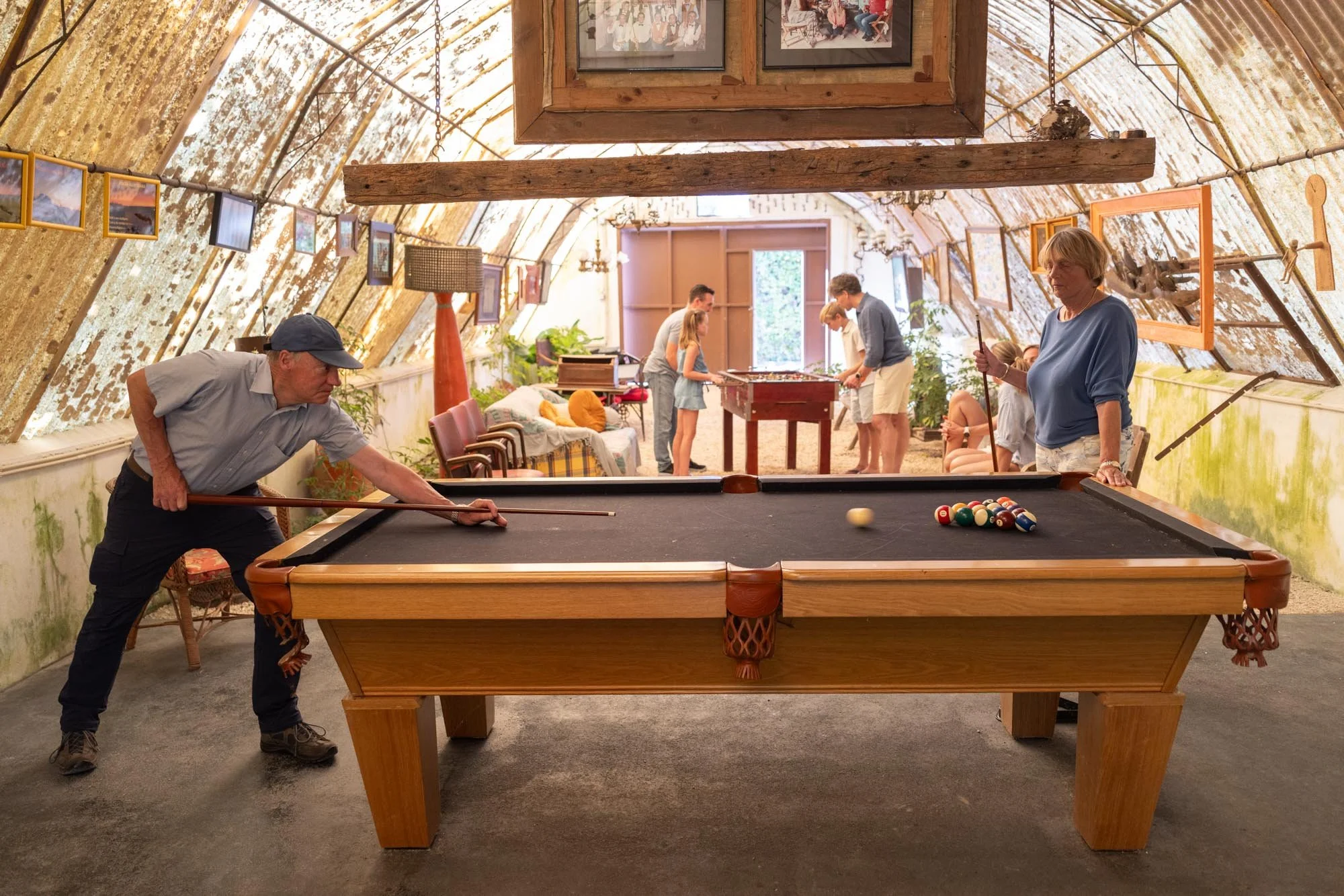 People playing billiards in an attic-style room with furniture and framed photos on the walls.