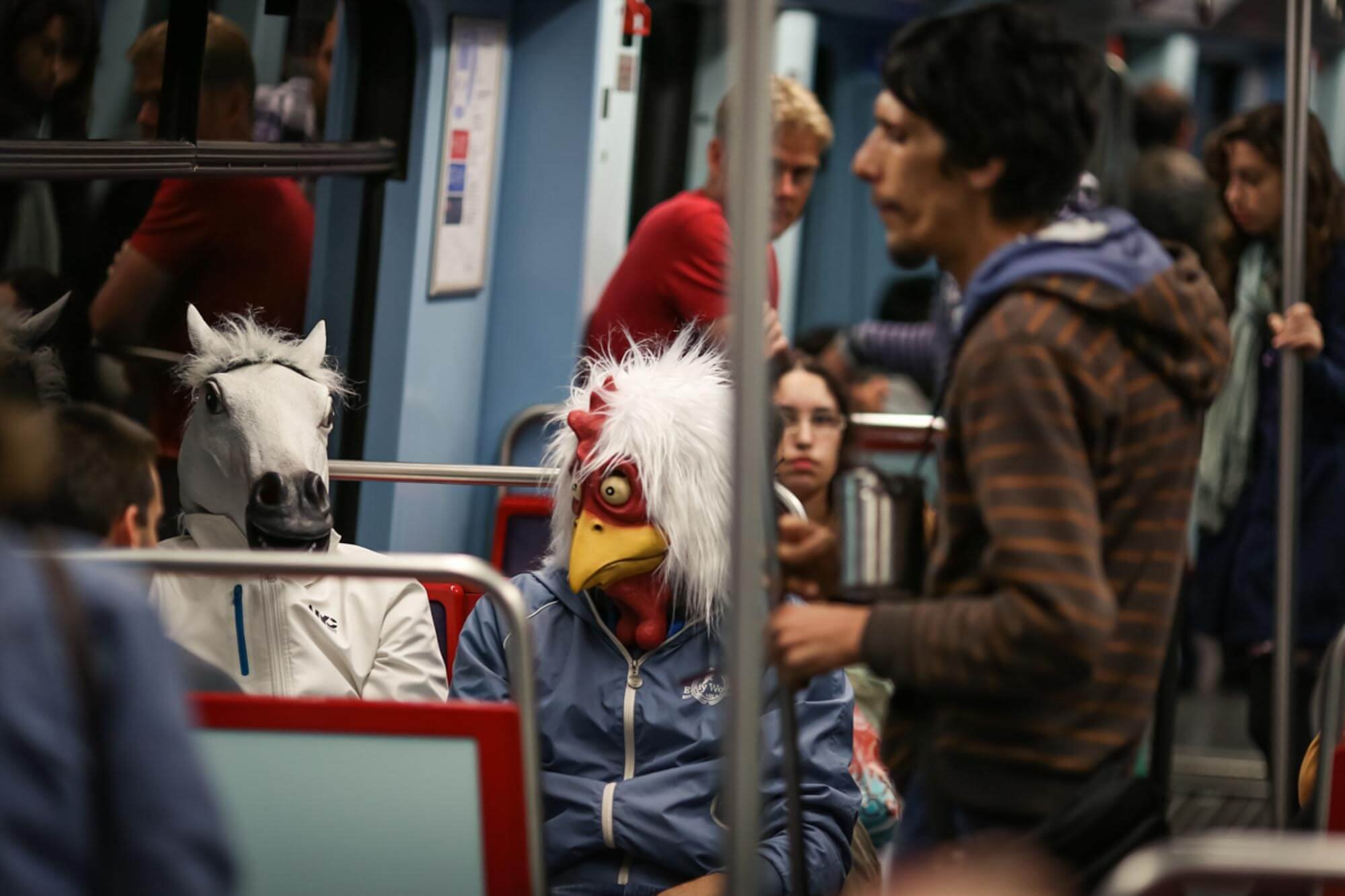 Two people wearing horse and rooster masks sitting on a bus, with a man standing nearby holding a drink and a woman in glasses in the background.