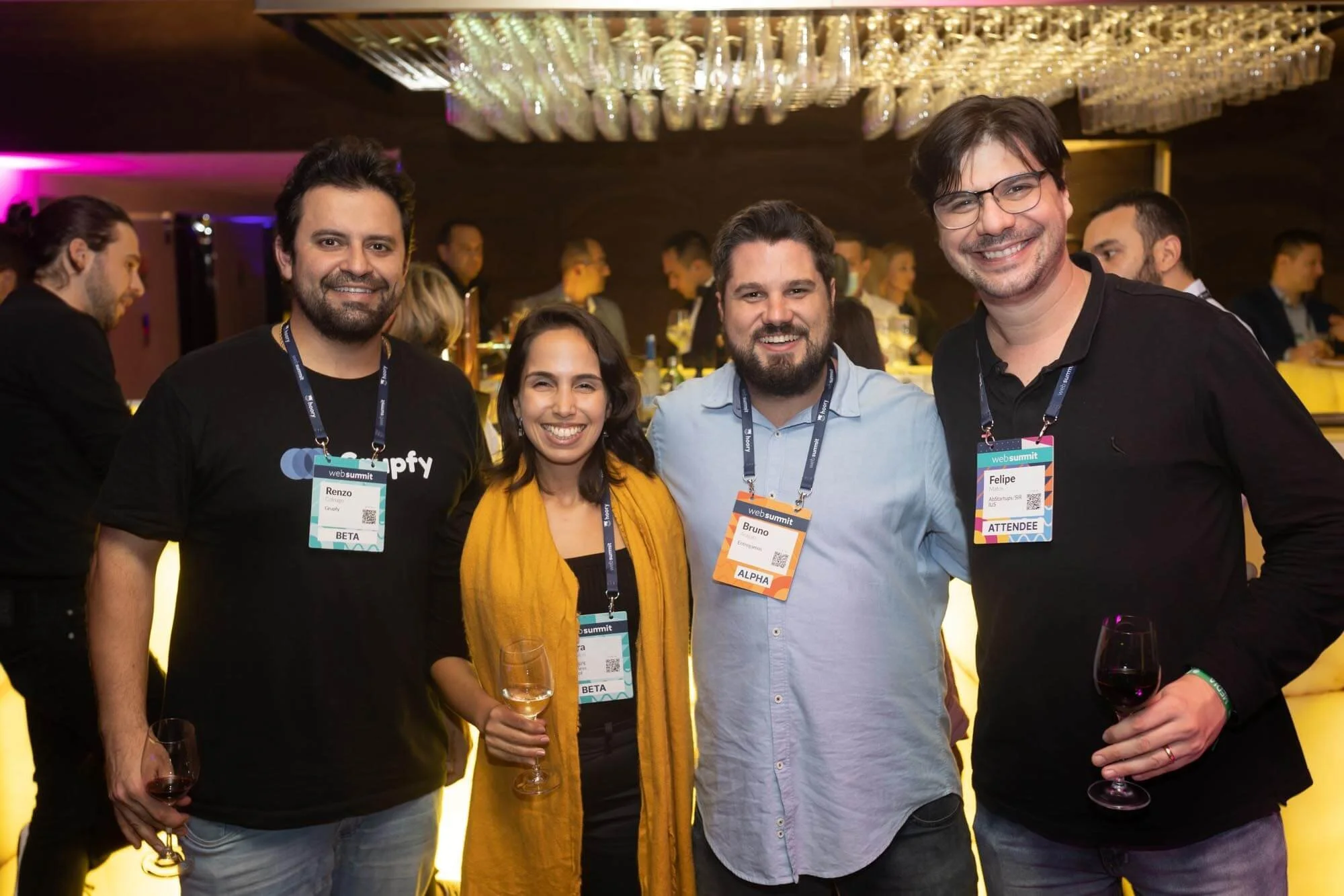 Four people smiling at a networking event, holding wine glasses, with name tags around their necks, in a dimly lit room with hanging glassware and other attendees in the background.