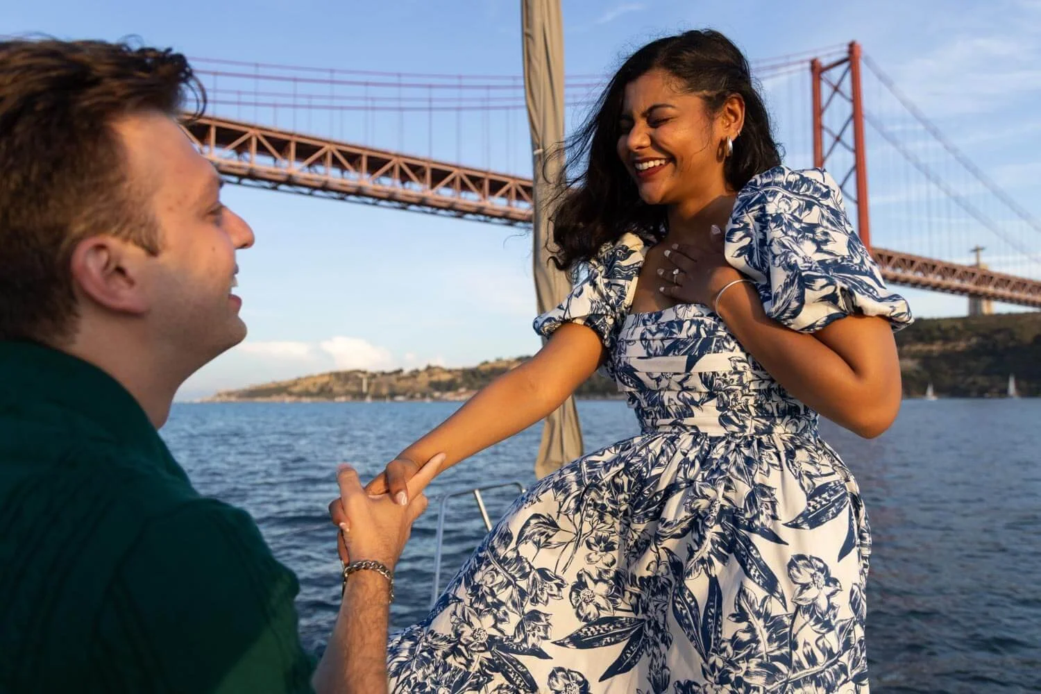 A man kneels on a boat holding a woman's hand, who is smiling and holding her chest, with the San Francisco Bay Bridge in the background during sunset.