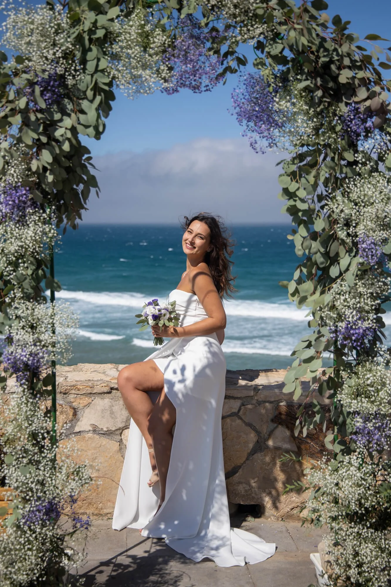 A woman in a white strapless dress sits on a stone ledge under a floral archway on a beach, holding a bouquet, with ocean waves and blue sky in the background.