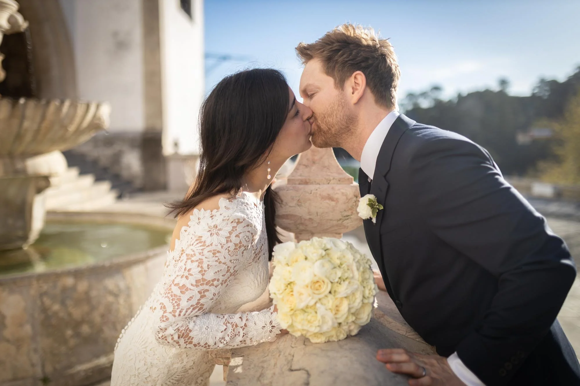 Bride and groom kissing outdoors with a fountain and stone railing in the background, the bride holding a bouquet of white flowers.