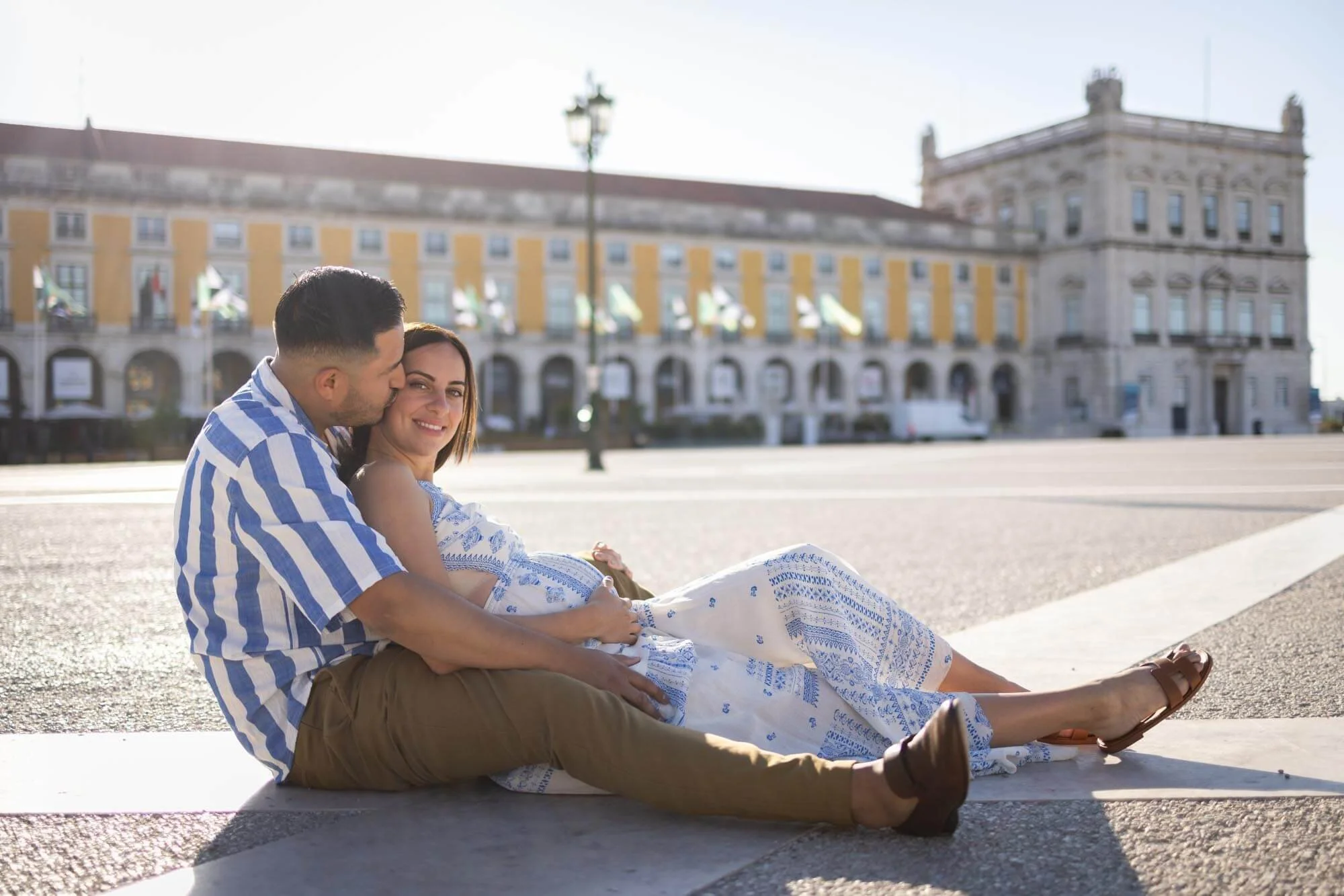 A couple sitting on the ground in a city square with historic buildings in the background. The man is kissing the woman's cheek, and she is smiling.