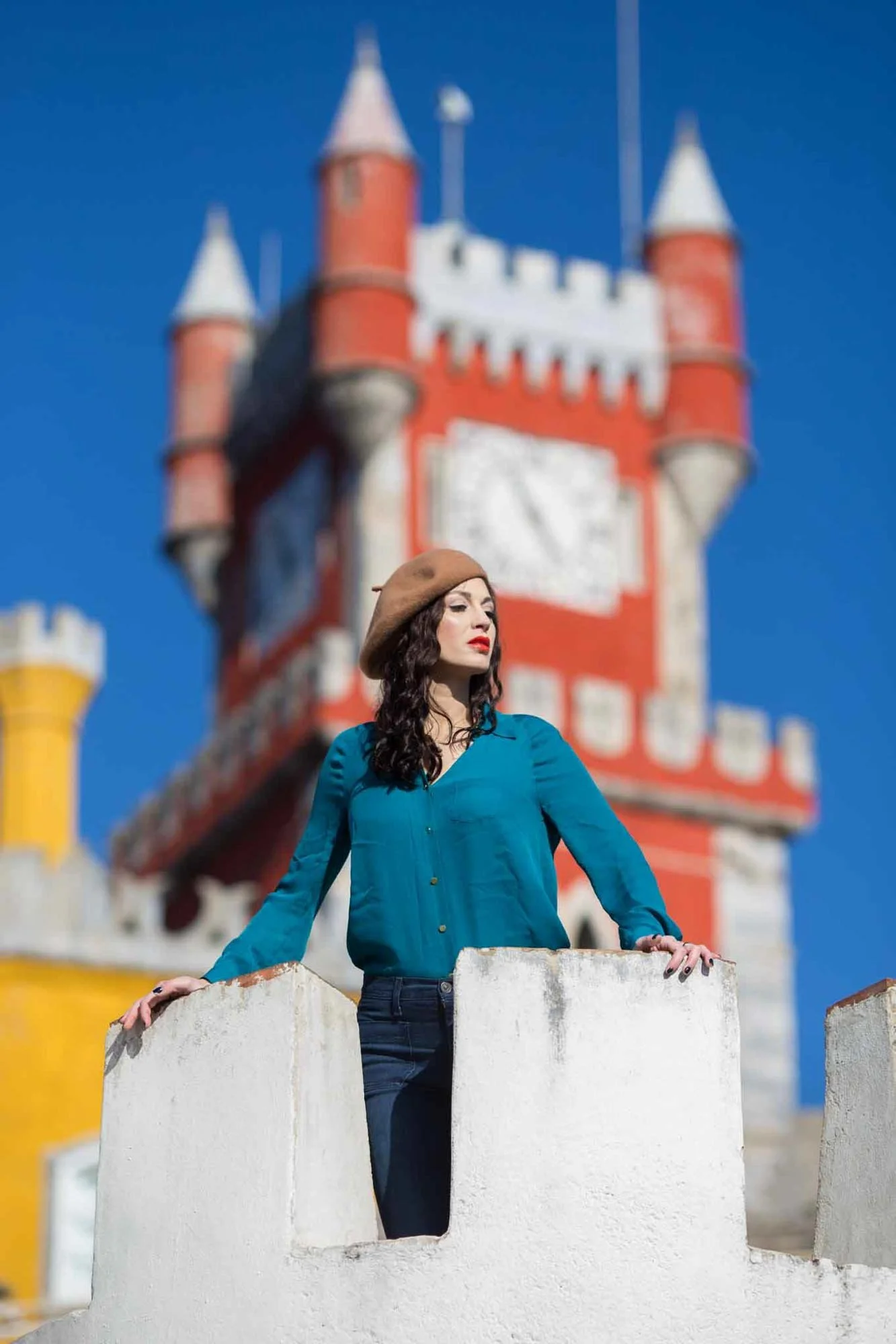A woman in a teal blouse and brown beret stands on a white wall, with a colorful castle in the background against a bright blue sky.