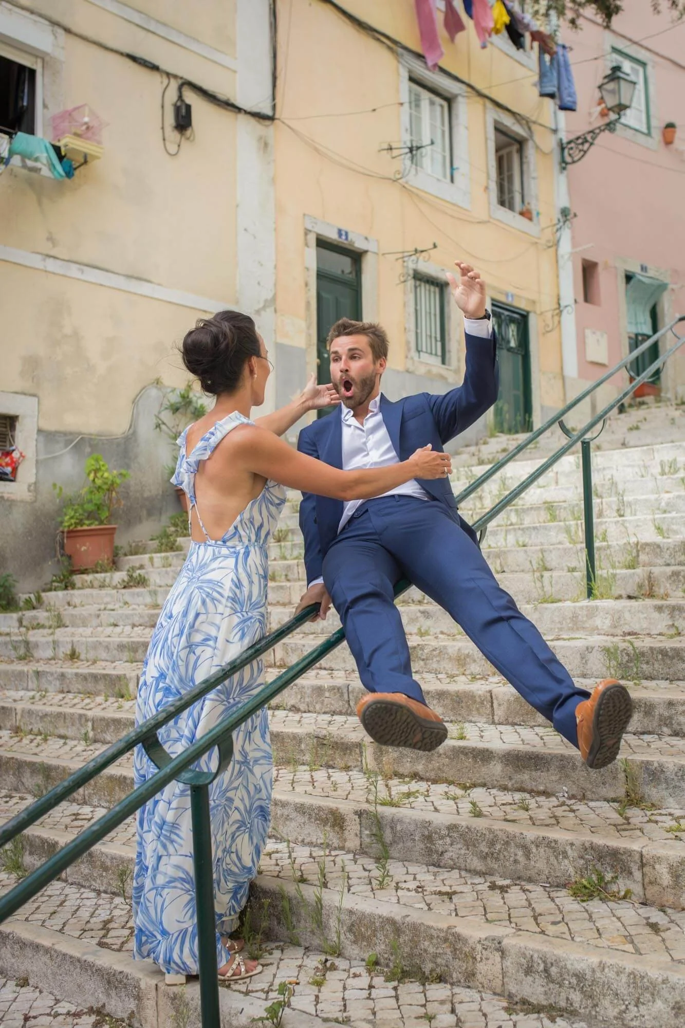 A woman in a blue and white floral dress appears to hit a man in a blue suit sitting on a stone staircase in a colorful, old European neighborhood.