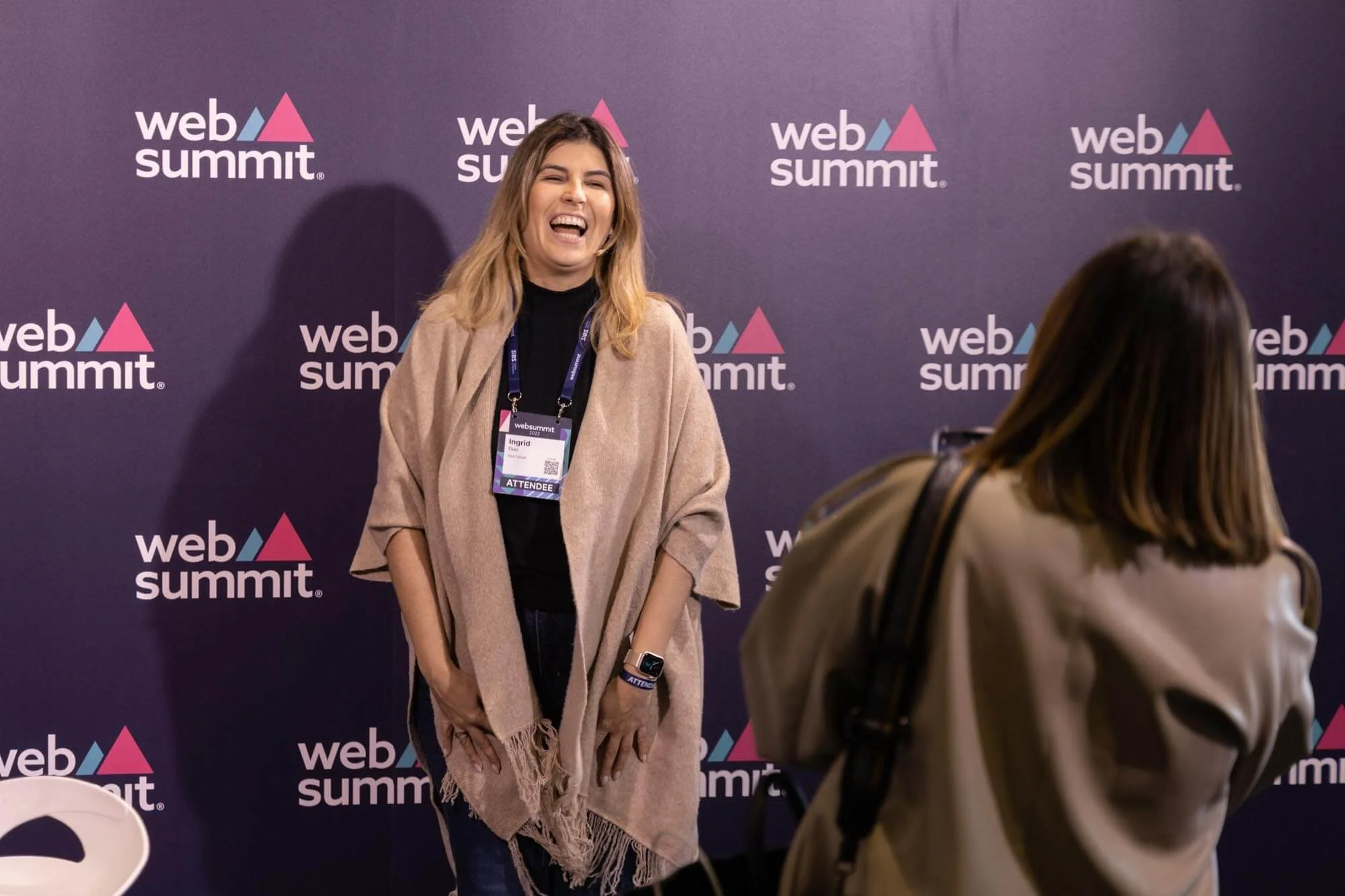 A woman at a web summit event, laughing and talking with another woman who has her back to the camera. The woman on the left wears a beige shawl, black top, and has a lanyard with an attendee badge. The background features a repeating 'web summit' lo