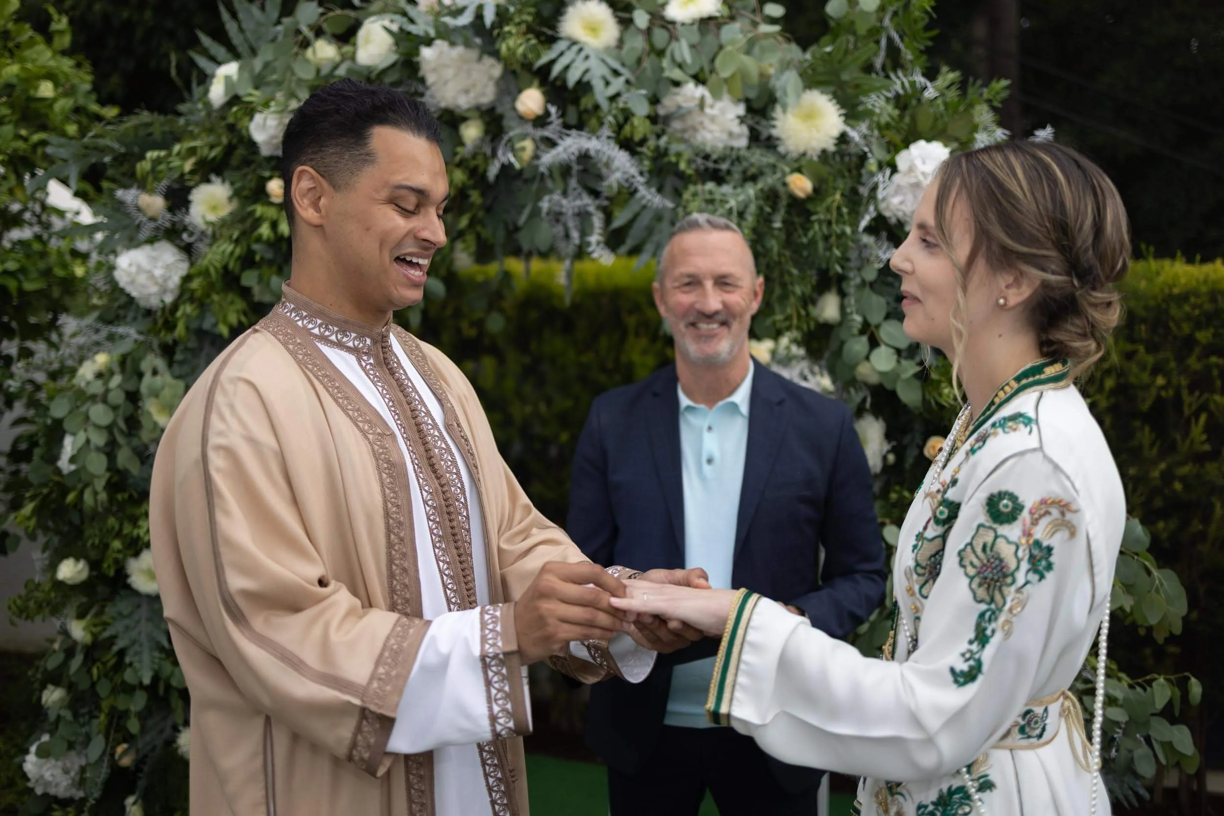 A couple exchanging rings during a wedding ceremony outdoors with a floral arch in the background, officiant smiling behind them.