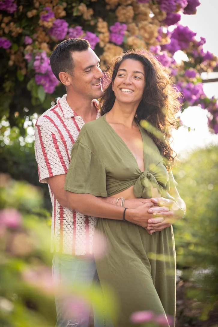 A couple standing outdoors among colorful flowers, with the man standing behind the woman, both smiling and looking at each other, during warm sunlight.