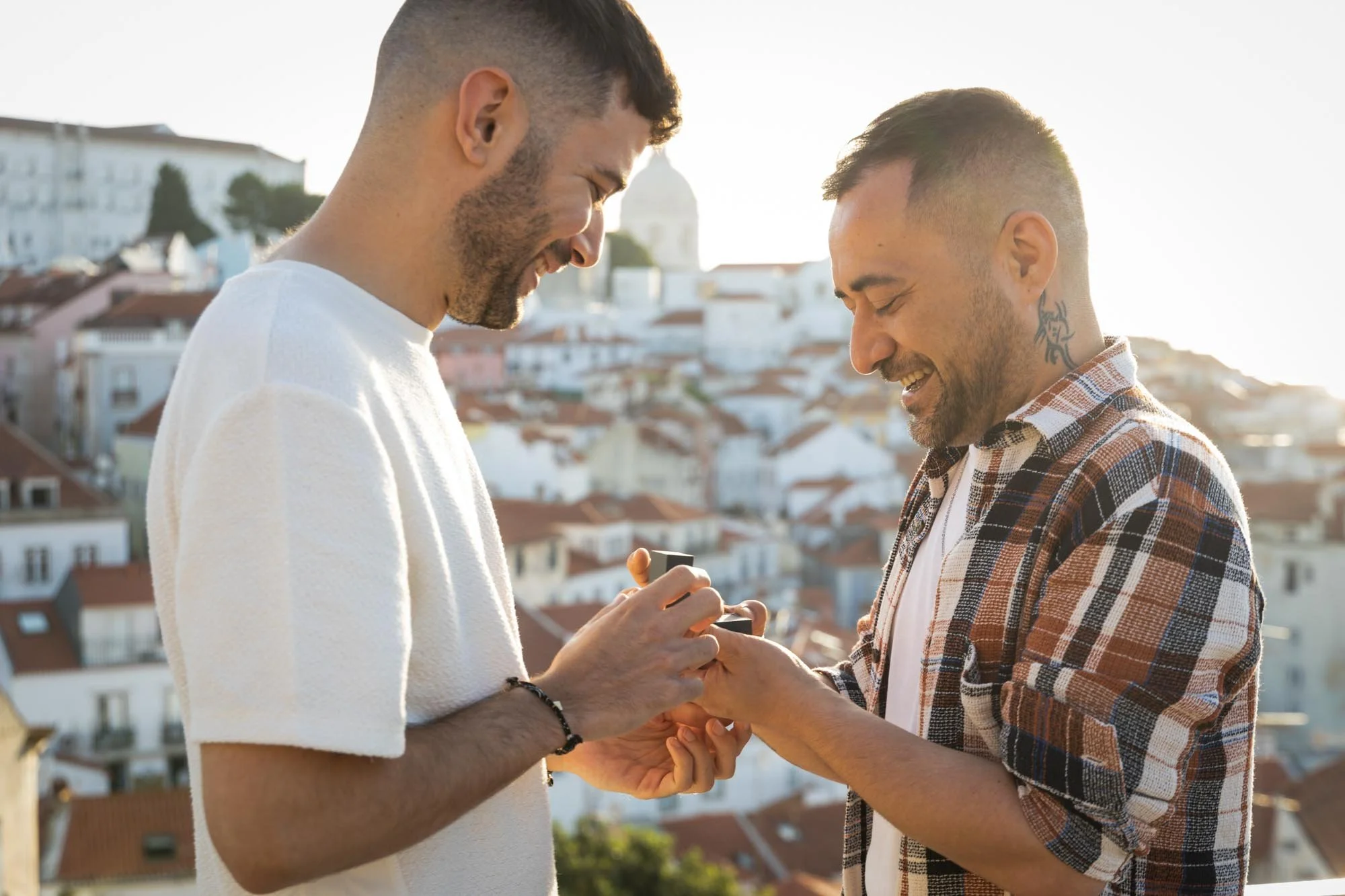 Two men are standing close together on a rooftop, smiling and looking at a smartphone. One man is in a white t-shirt, and the other is in a plaid shirt. The background shows a cityscape with houses and a domed building in the distance, under a bright