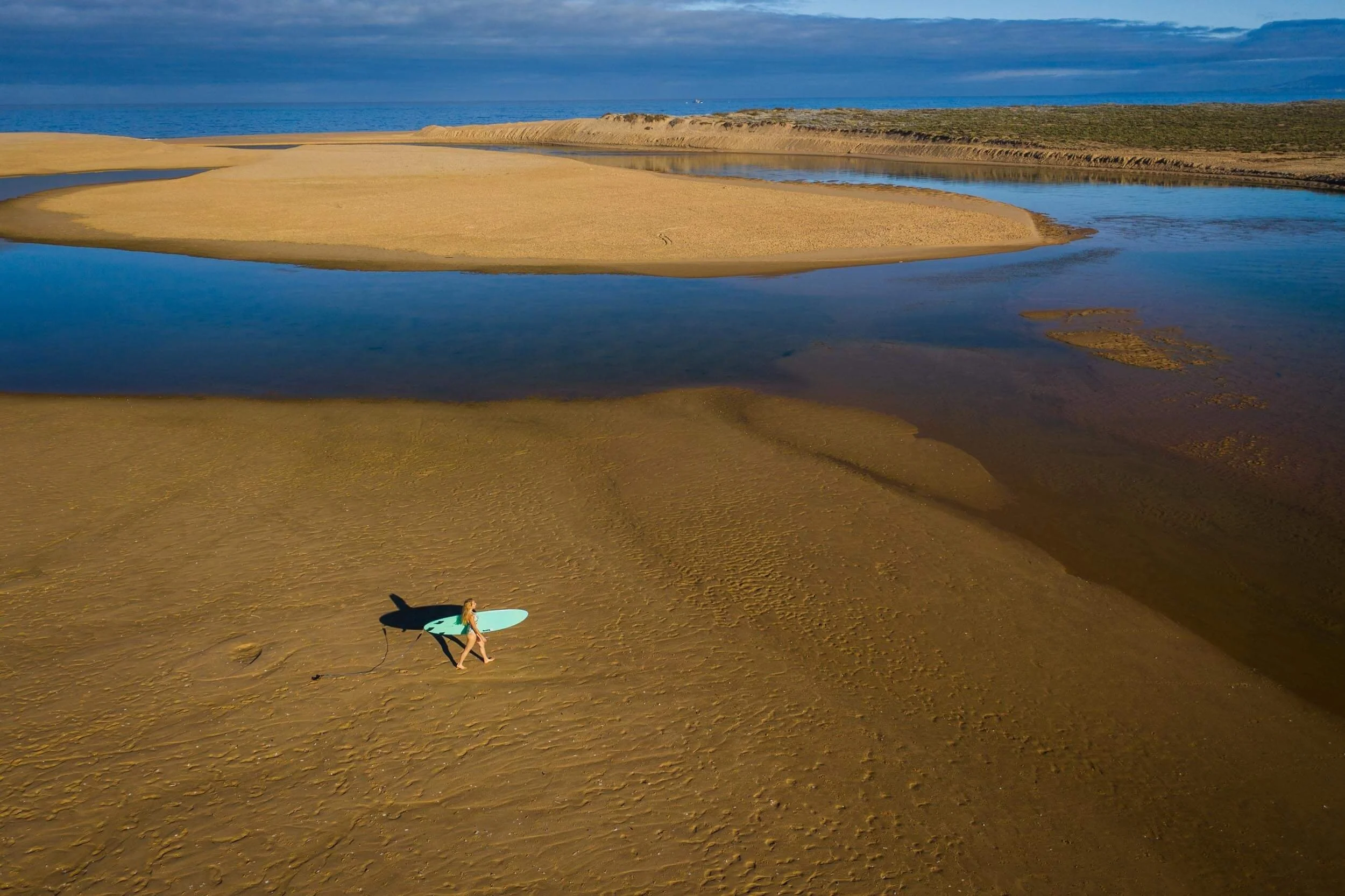 A woman carrying a surfboard walking on sandy beach with tidal pools and ocean in the background.