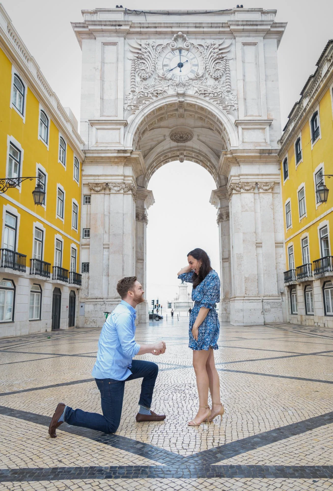 A man proposes to a woman in front of Arco da Rua Augusta in Lisbon, Portugal, on a cobblestone square with yellow buildings.