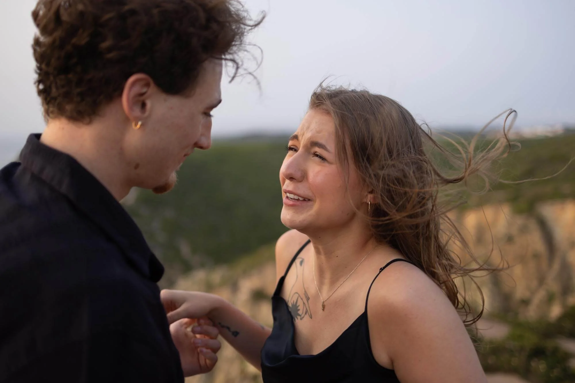 A young woman with long hair and a tattoo on her shoulder is crying while looking up at a young man with short curly hair. They are outdoors on a windy day with green hills in the background.