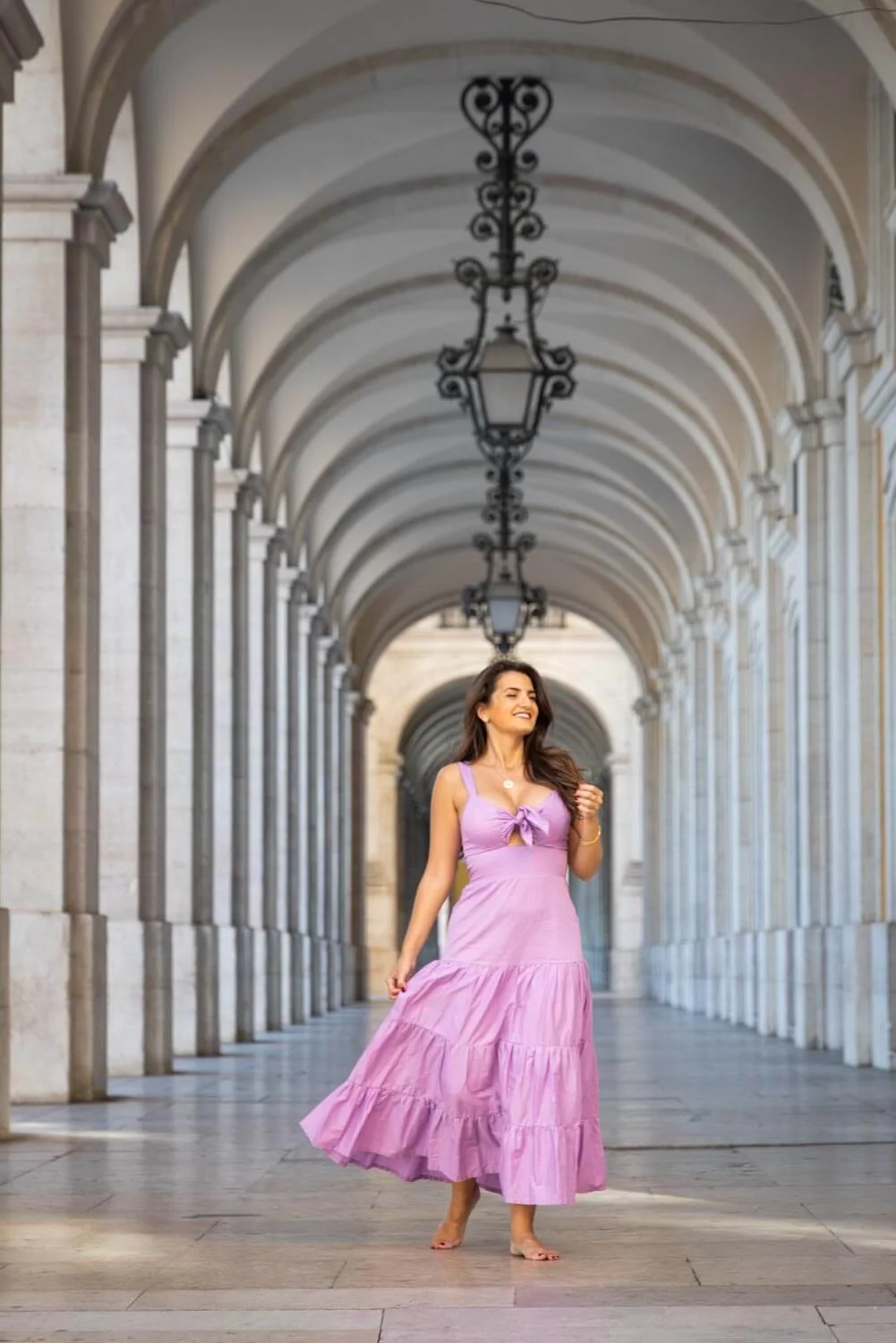 A woman in a pink dress walking barefoot through an ornate, arched corridor with hanging lanterns.