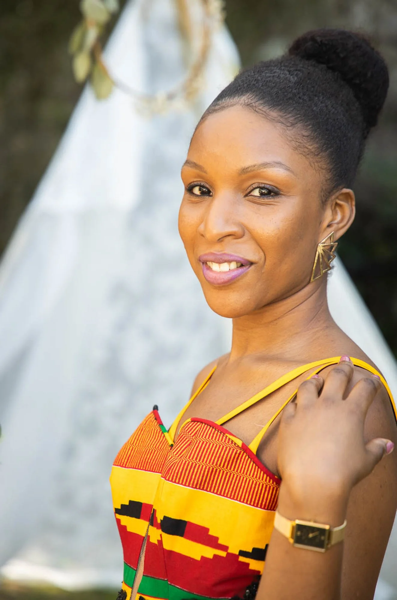 Portrait of a woman with dark hair in a bun, wearing a sleeveless vibrant dress with a red, yellow, black, and green pattern, gold earrings, and a gold watch, standing outdoors with a white dress hanging in the background.