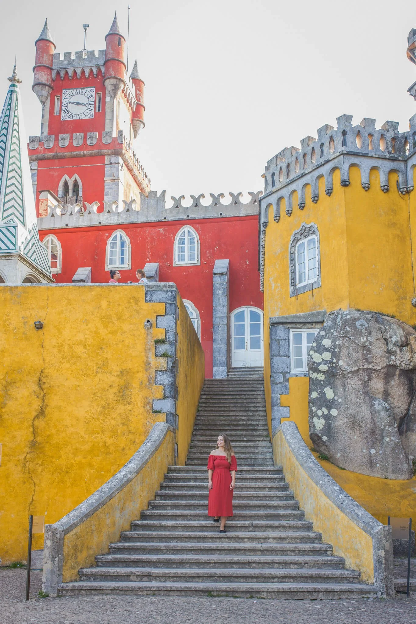 A woman in a red dress walking down stone stairs in front of a colorful, castle-like building with yellow and red walls, arched windows, and a clock tower.