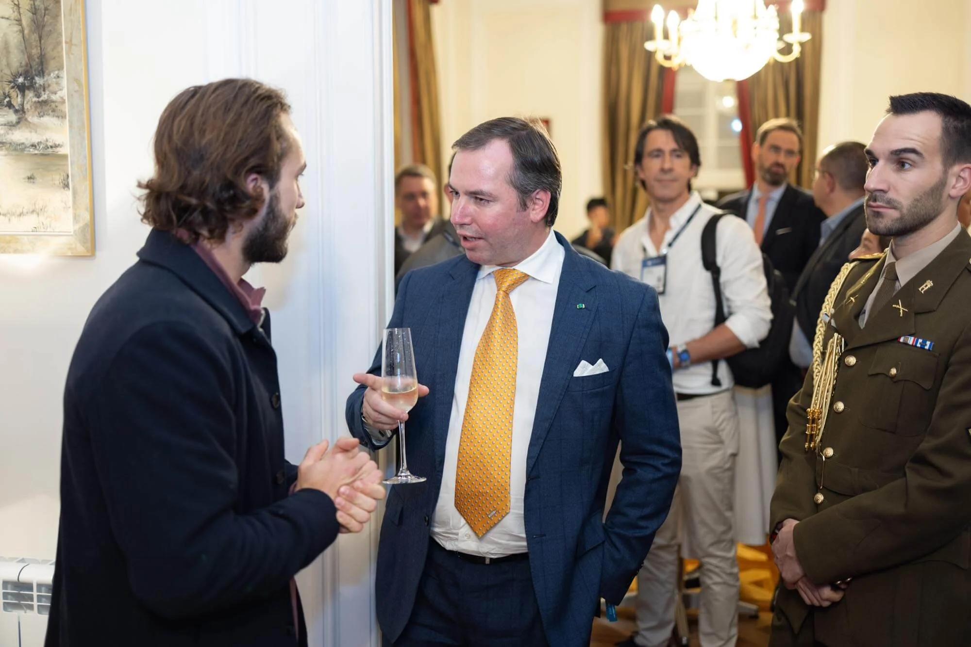 Three men having a conversation at a social event, with one holding a glass of champagne, in an elegant room with a chandelier and painting on the wall.