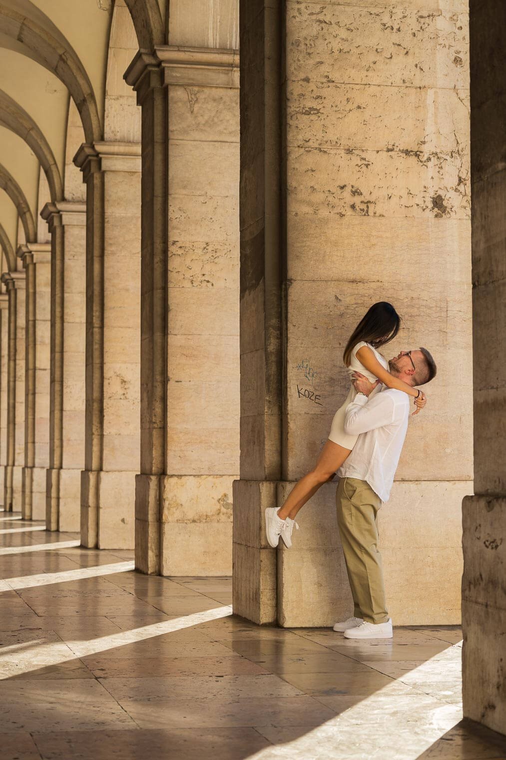 A man lifting a woman against a stone wall in a colonnade with arches and sunlight casting striped shadows on the floor.