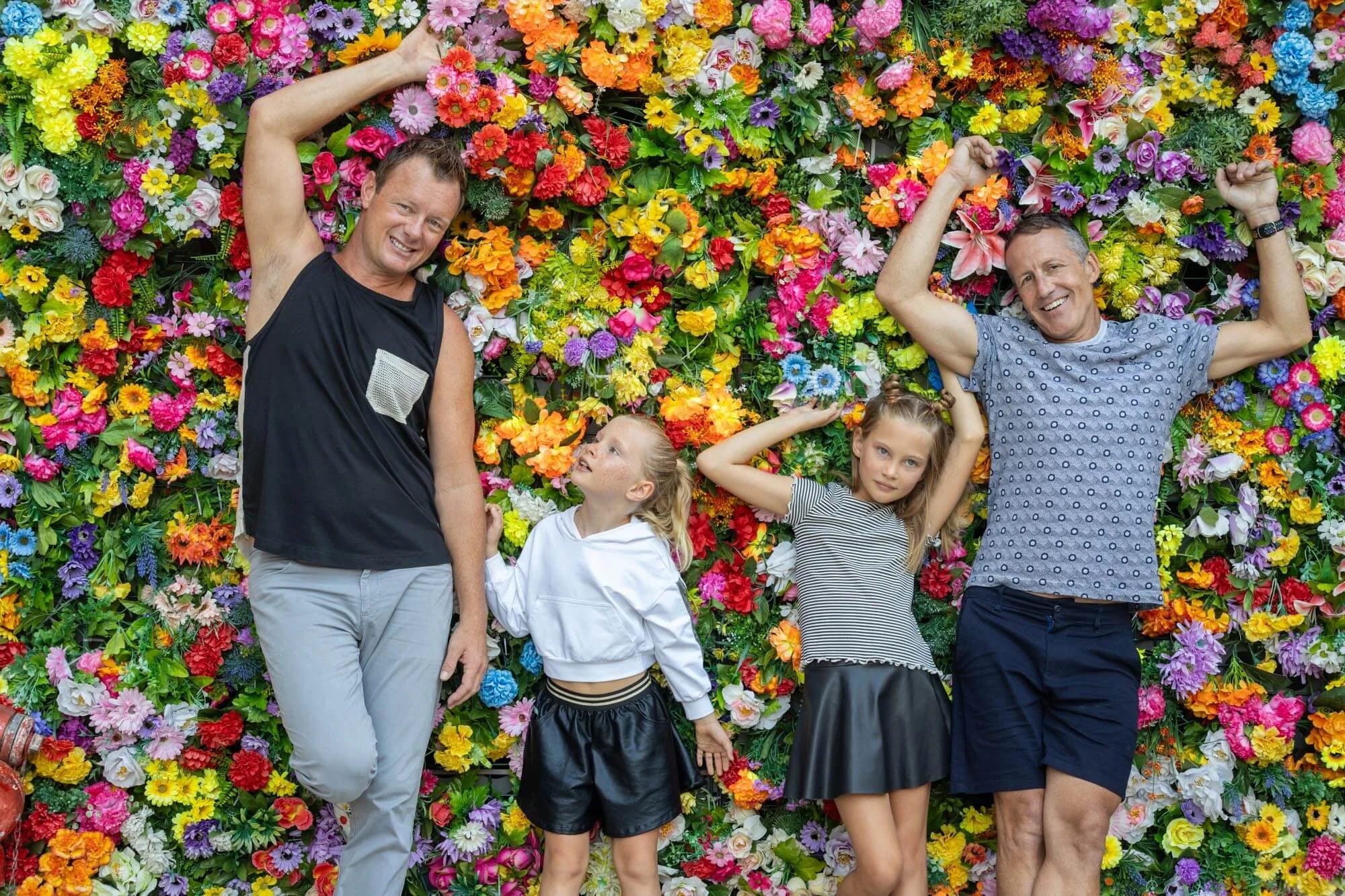 A family of four, two adult men and two young girls, lying on a colorful wall of flowers, smiling and looking happy.