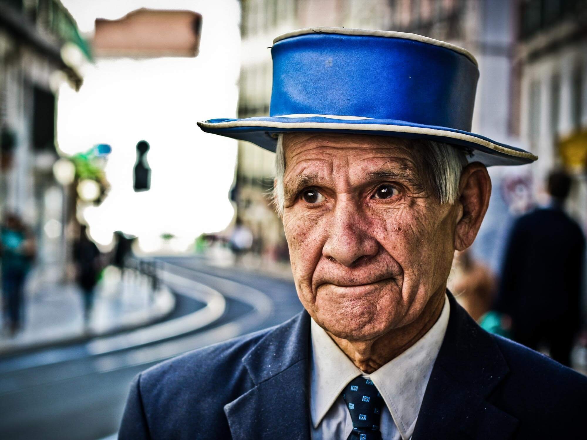 An elderly man wearing a blue top hat and a dark suit, standing outdoors in a city street.