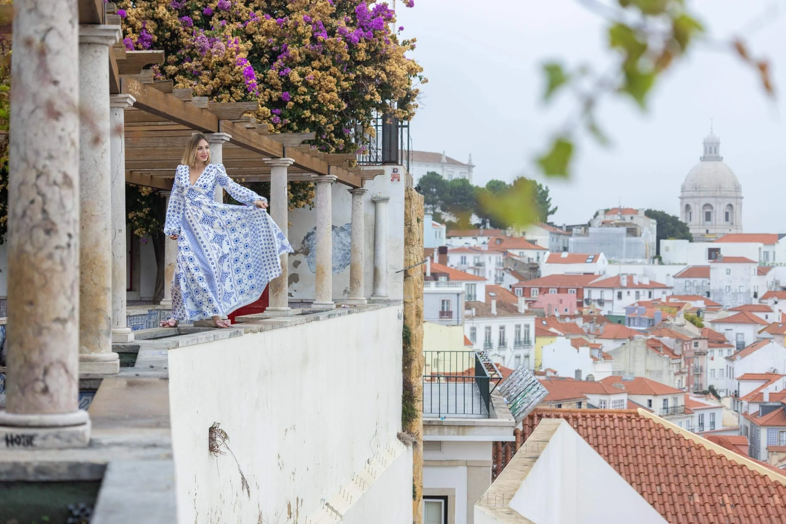 A woman in a blue and white patterned dress stands on a balcony overlooking a cityscape with white buildings and red-tiled roofs, and a large domed building in the background, with some greenery and blooming flowers nearby.