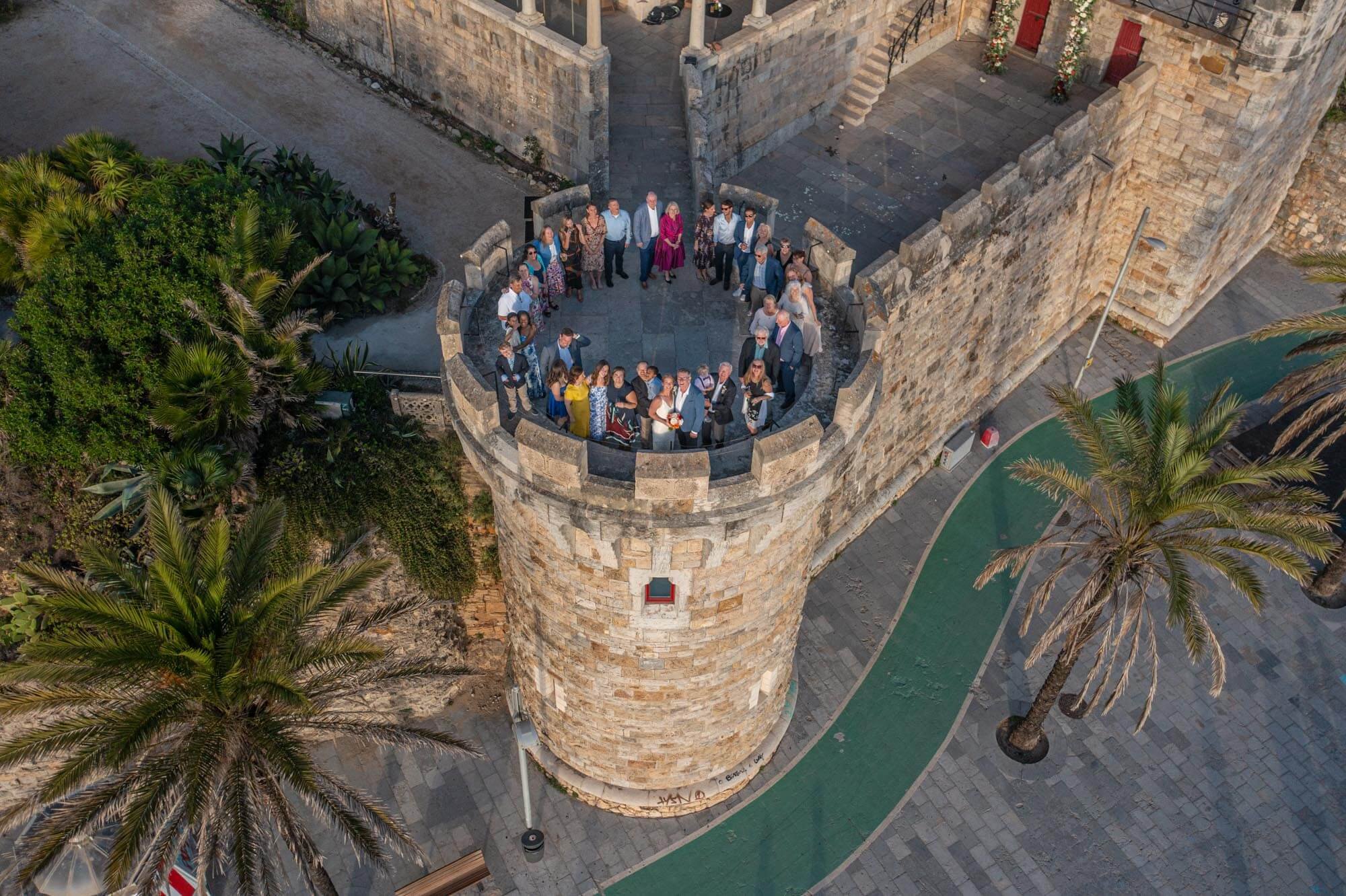 Aerial view of a group of people gathered on a stone tower of a historic castle or fortress, with palm trees and a paved walkway below.