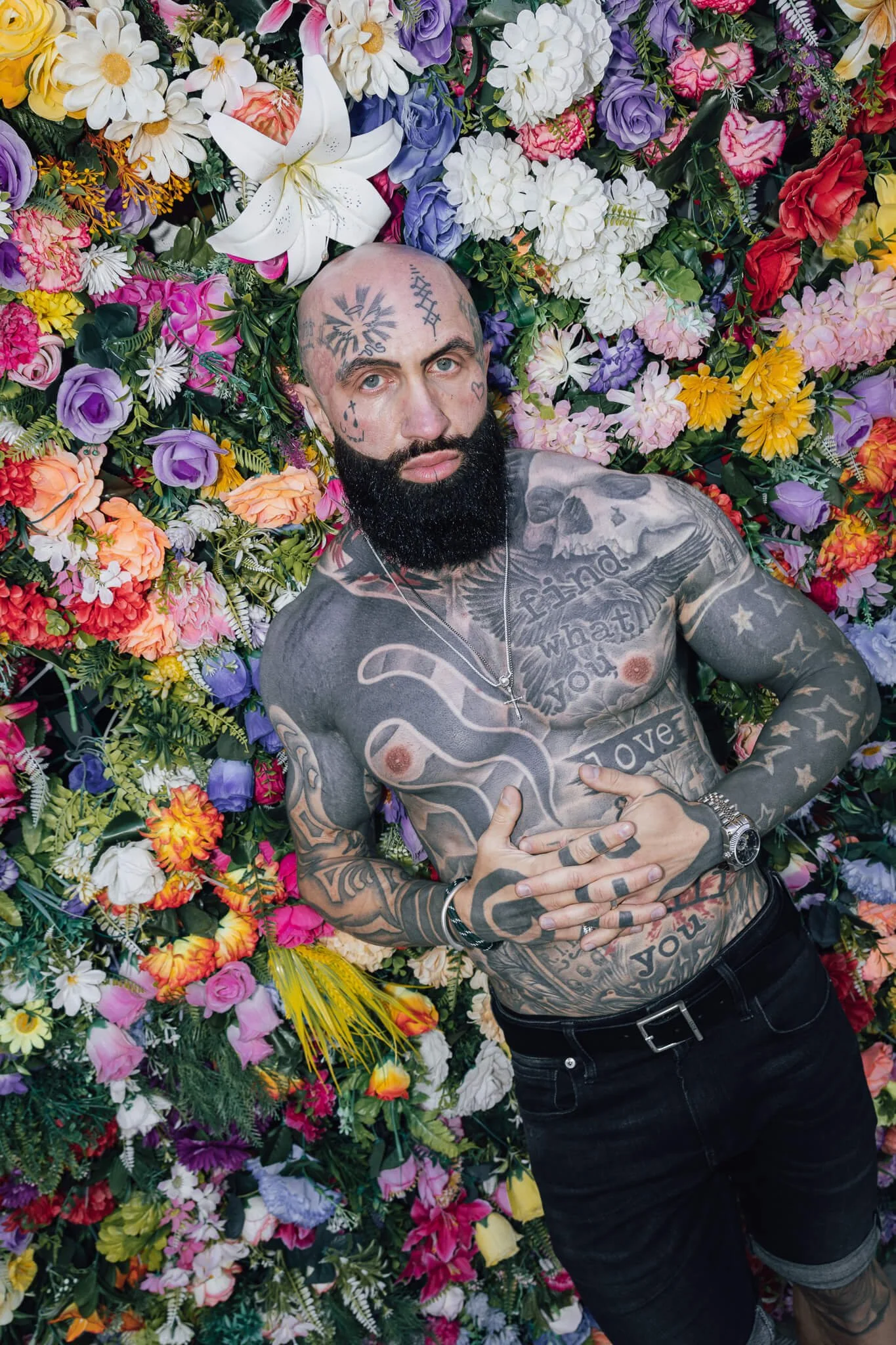 A tattooed man with a beard lying on a colorful flower wall, wearing black shorts, multiple rings, a watch, and a necklace.